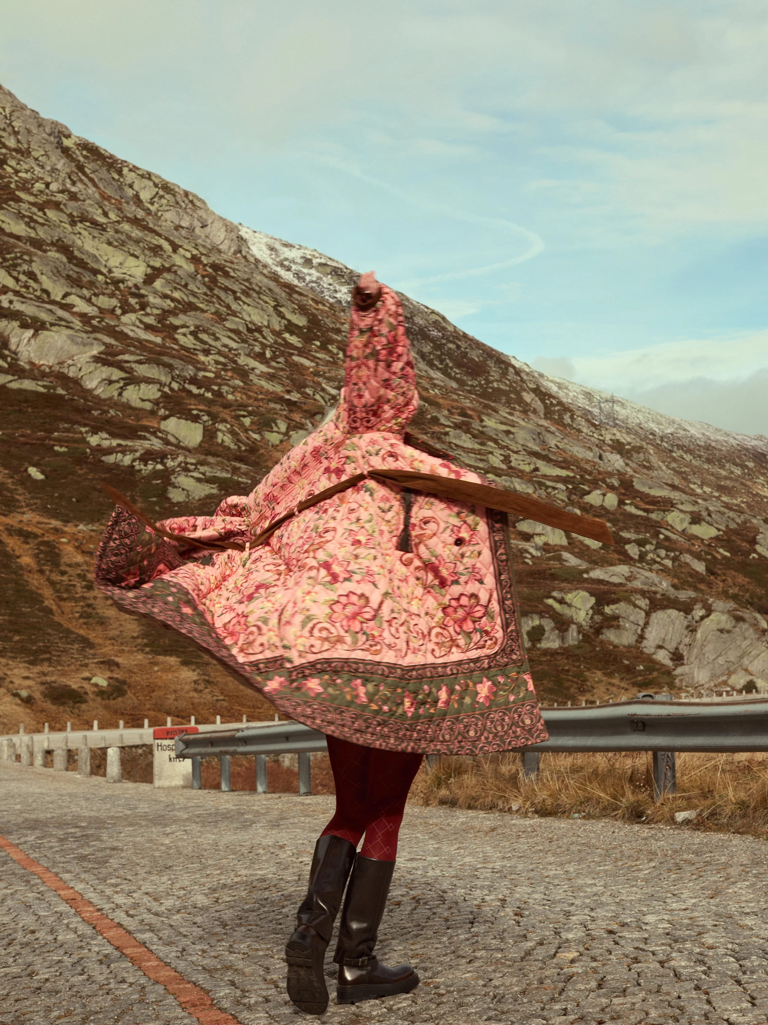 Ein Mensch mit einem Kleid in Blumendesign, der auf einer geteerten Straße in bergigem Gelände steht.