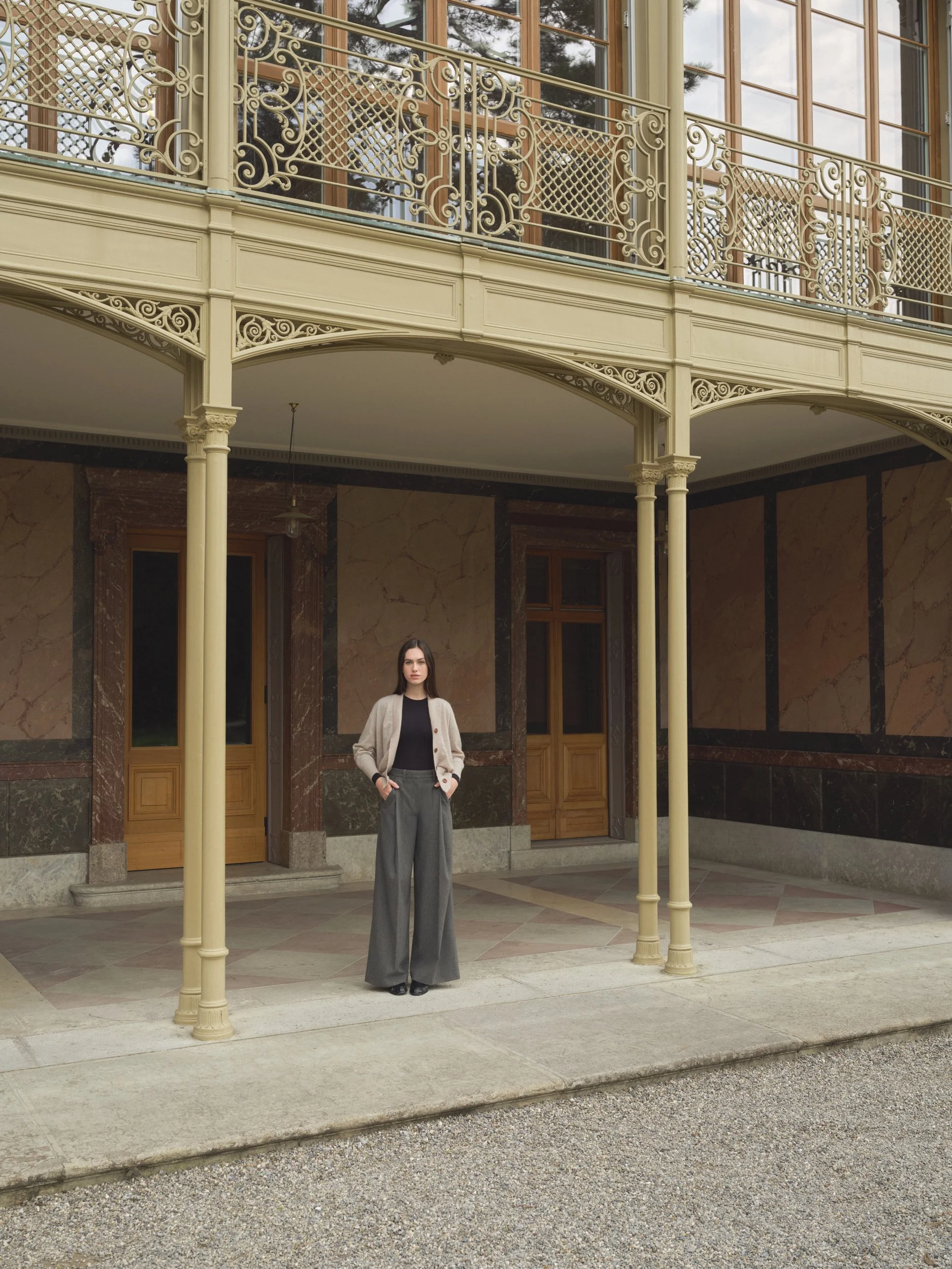 Junge Frau in eleganten Hosen und Cardigan, stehend vor einem historischen Gebäude mit viel verzierten Metallgeländer und großen Fenstern.