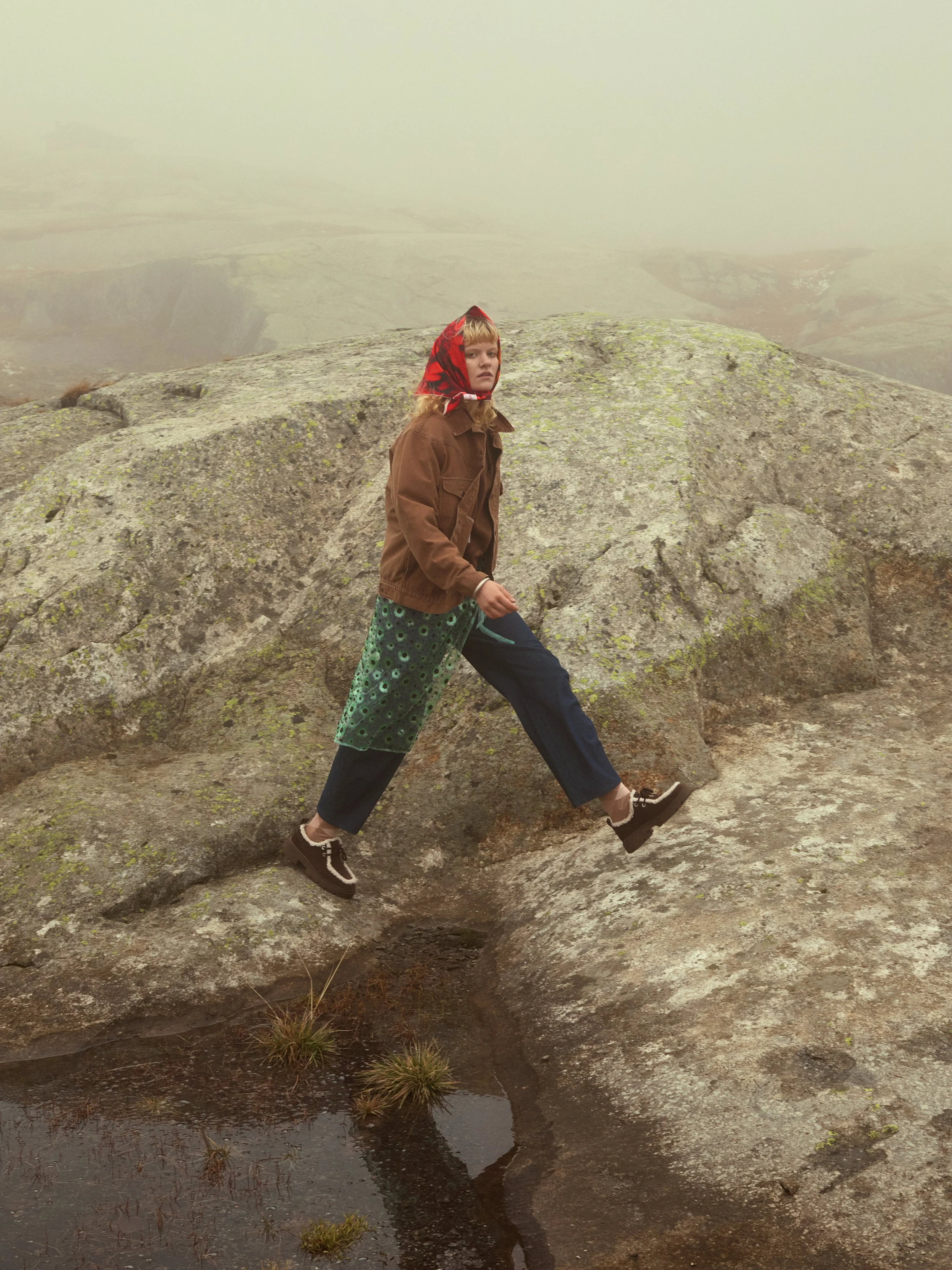 Ein Mädchen in Outdoor-Kleidung und einem roten Kopftuch, das auf einem großen, moosbedeckten Felsen an einem nebligen Tag auf einer felsigen Landschaft steht.