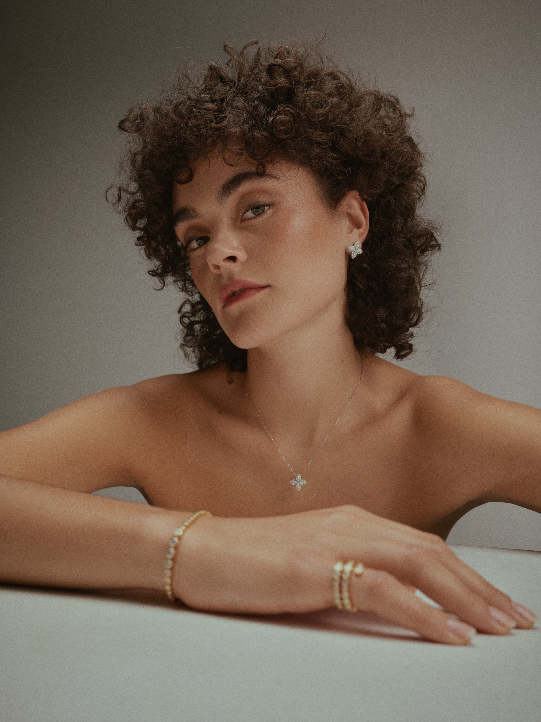 Nahaufnahme einer jungen Frau mit lockigem Haar, die Schmuck trägt, darunter Ohrringe, eine Halskette, ein Armband und eine Perlenkette, vor neutralem Hintergrund.