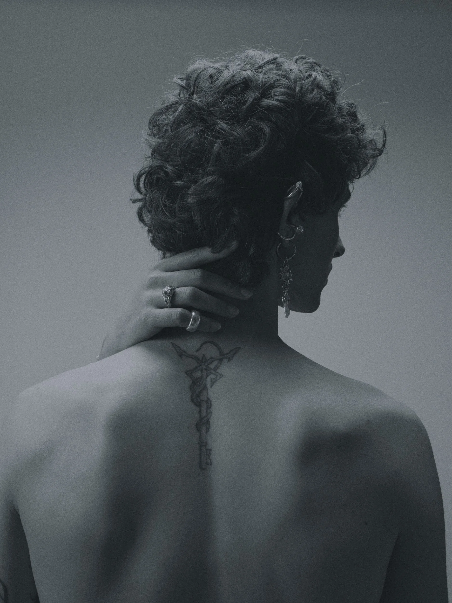 Schwarz-Weiß-Fotografie eines männlichen Models mit kurzen, lockigen Haaren, mehreren Ohrringen, einer Hand mit mehreren Ringen, einer Halskette mit Anhänger, einer Tattoo am Hals, der Kopf nach rechts geneigt.