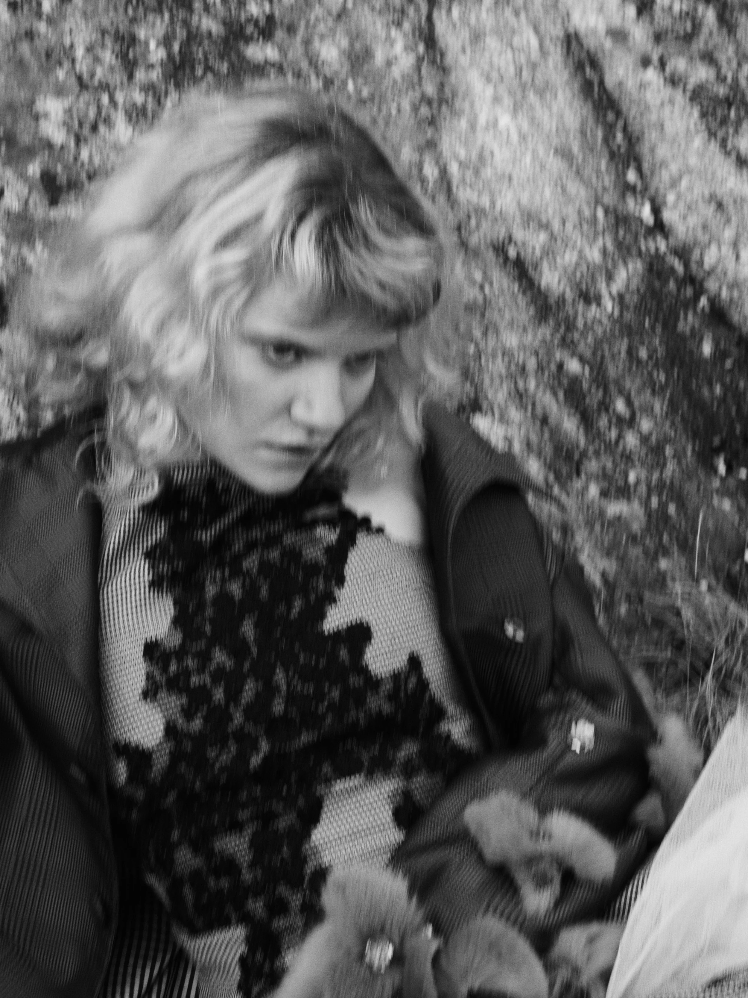 Schwarz-Weiß-Foto einer jungen Frau mit lockigem, mittellangem Haar, die auf dem Boden liegt und in die Kamera schaut, mit einem metallischen, durchbrochenen Top und einem dunklen Jacke, vor eine felsige Wand.