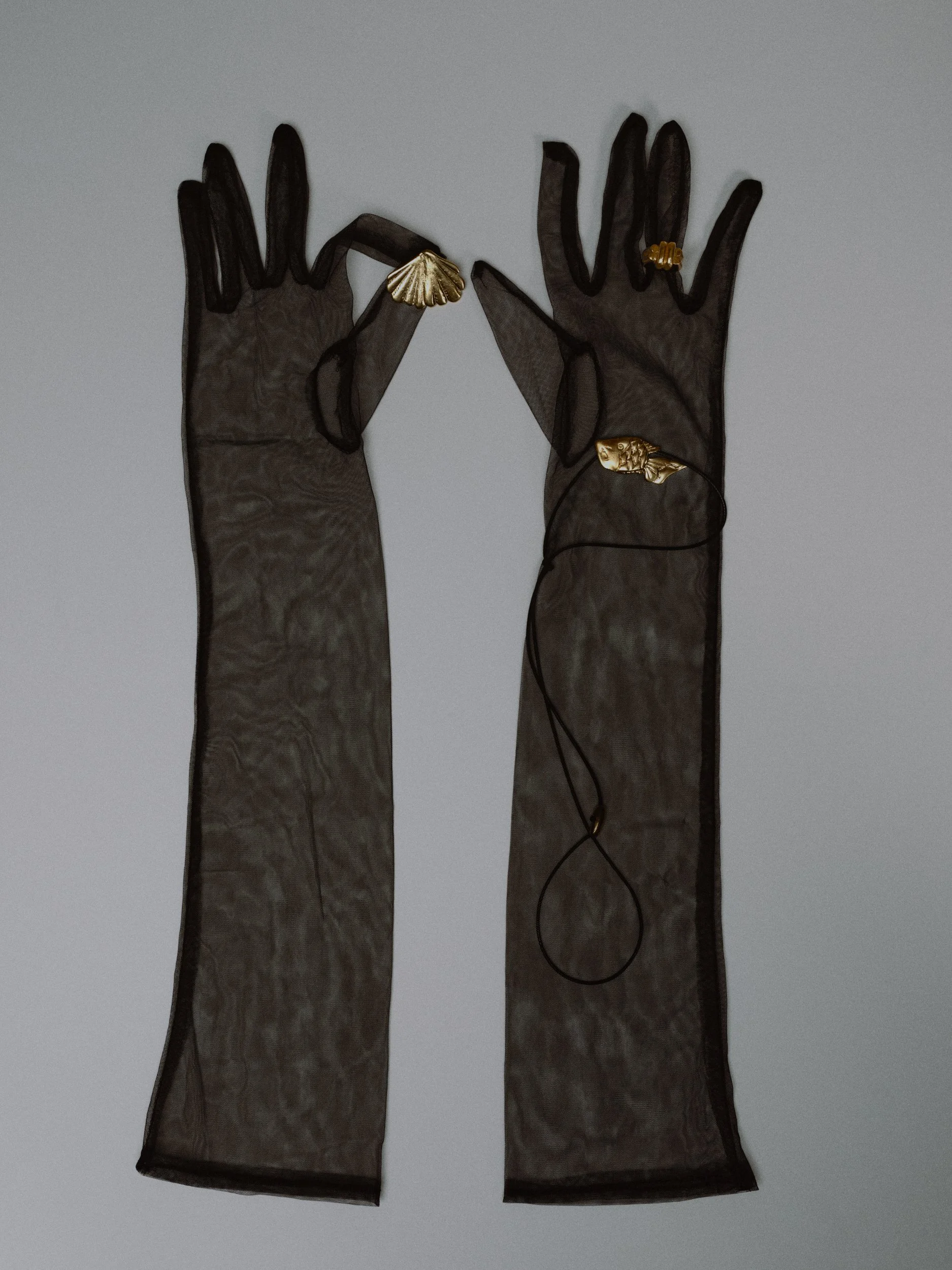 Zwei schwarze, transparente Handschuhe mit goldenen Schmuckstücken, einer in Form einer Muschel, der andere in Form eines Tierkopfes, auf einem weißen Hintergrund.