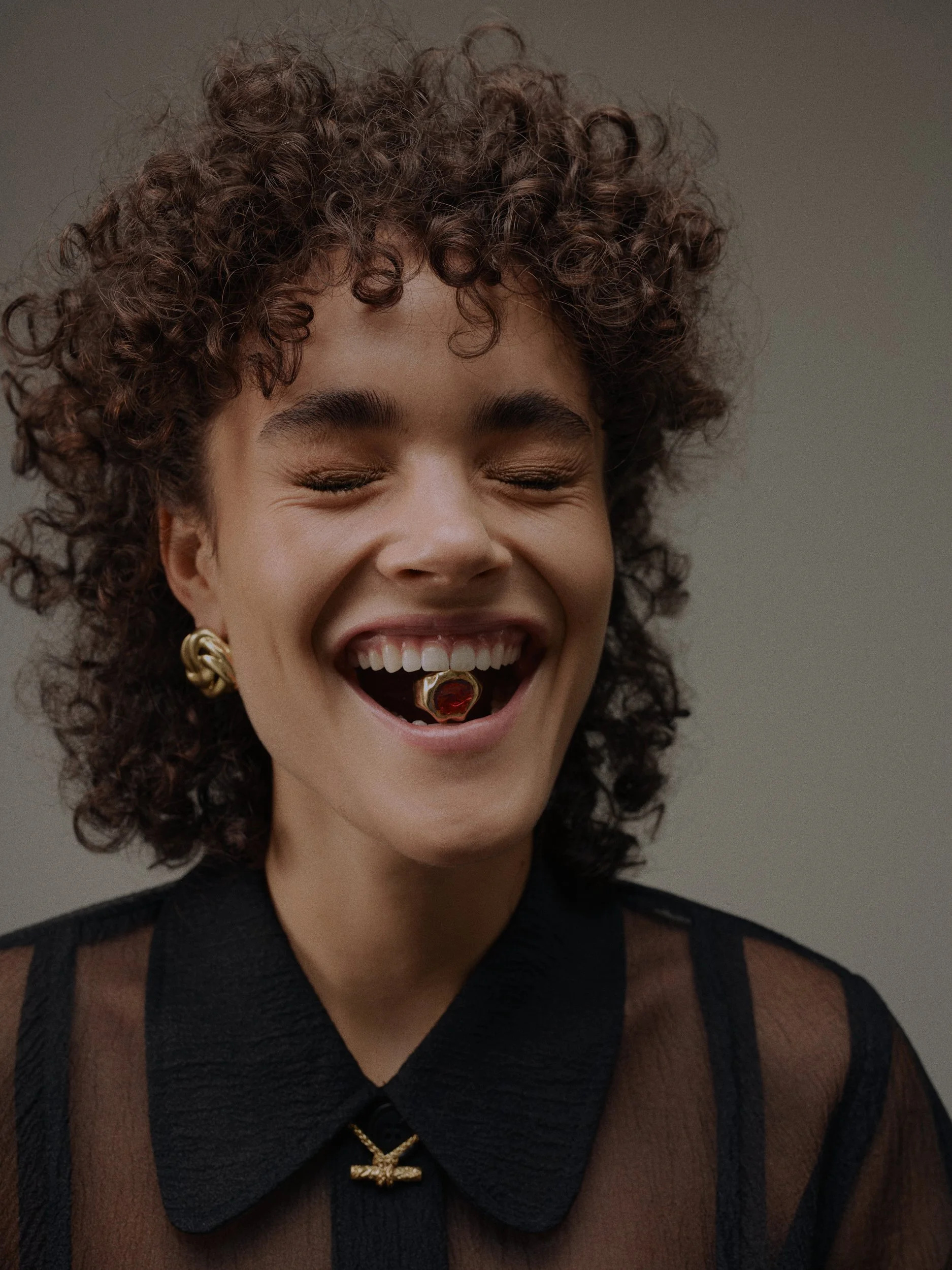 Ein weibliches Model mit kurzen, lockigen braunen Haaren, das lacht, mit Goldschmuck und einem dunkel durchscheinenden Kleid.