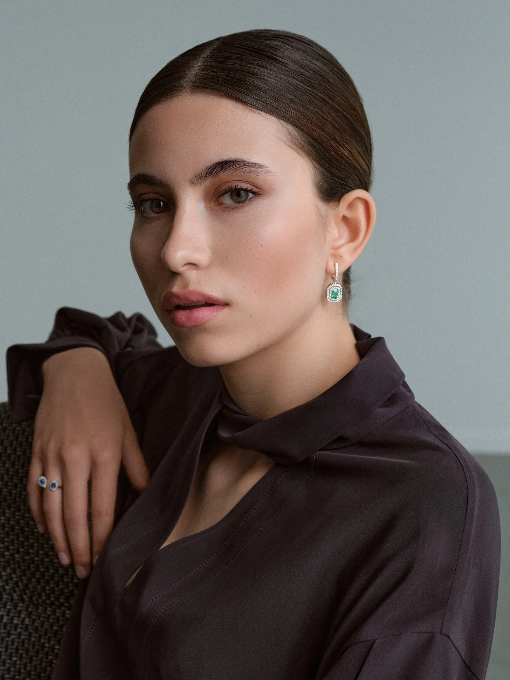 Porträt einer jungen Frau mit dunklem Kleid, die einen grünen Edelstein-Ohrring trägt und an einem schwarzen Ring mit blauen Steinen auf ihrer Hand zeigt.