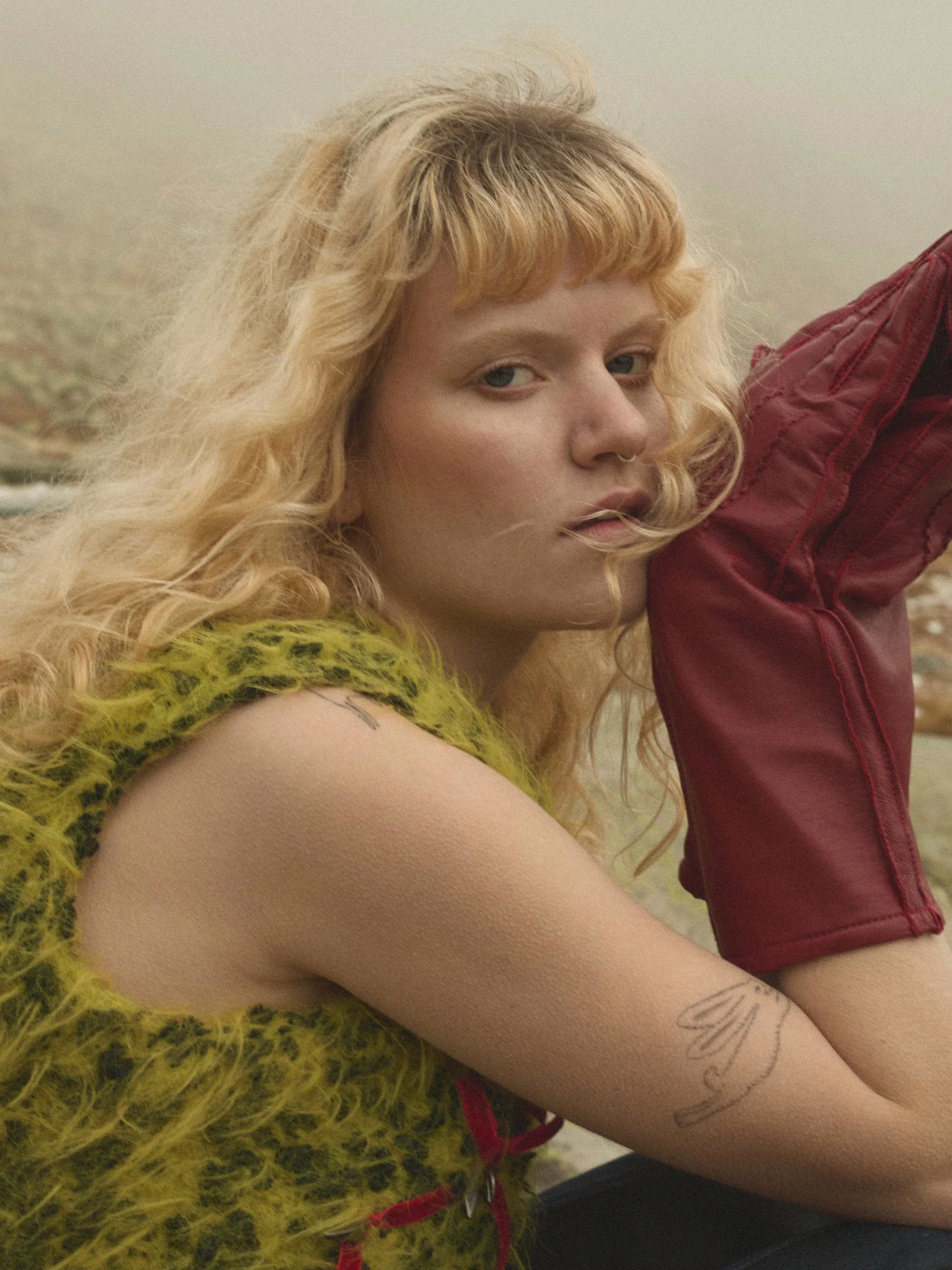 Eine junge Frau mit lockigem blondem Haar, die einen grünen, wolligen Pullover trägt, sitzt im Freien und lehnt ihren Arm auf ihr Knie, mit einem roten Rucksack. Sie blickt nach vorne, Neben dem Meer oder See, im Hintergrund neblige Landschaft.