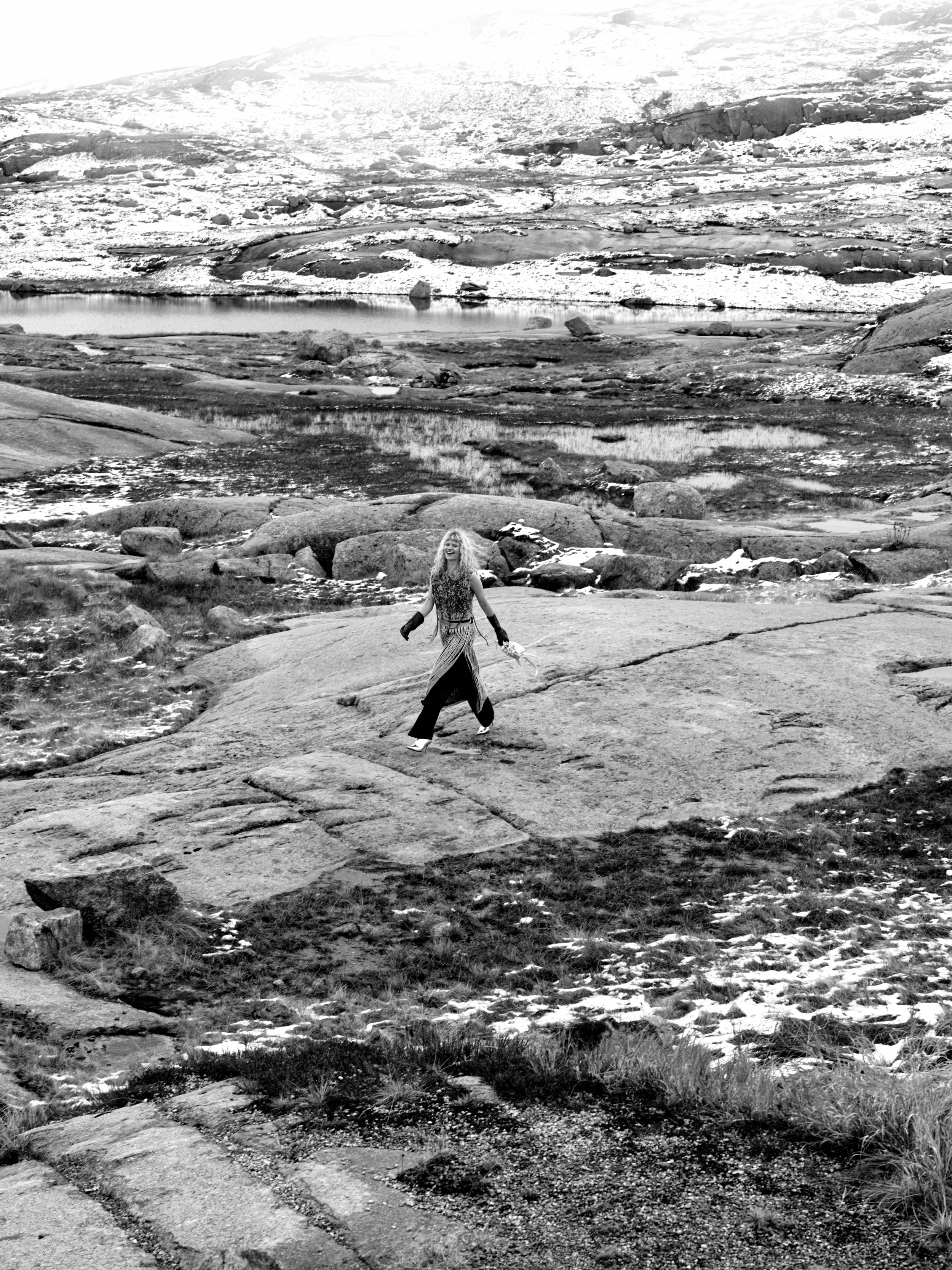 Eine Frau wandert auf einem Felsen in einer alpinen Landschaft mit Schnee, Wasser und Steinen in der Umgebung.