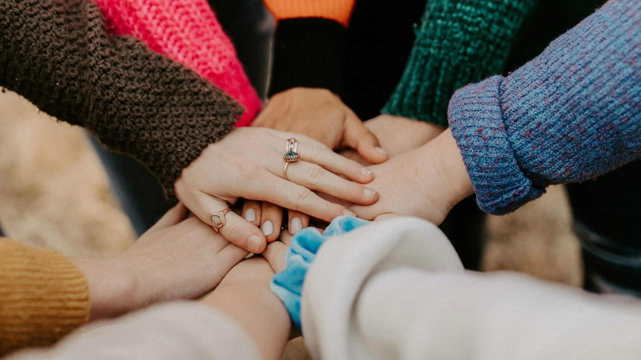 Multiple hands stacked together in a show of unity and support, with some hands wearing rings and others with colorful clothing.