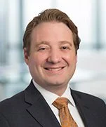 Headshot of a smiling man with light brown hair, wearing a dark suit and a gold patterned tie, in an office setting with large windows.