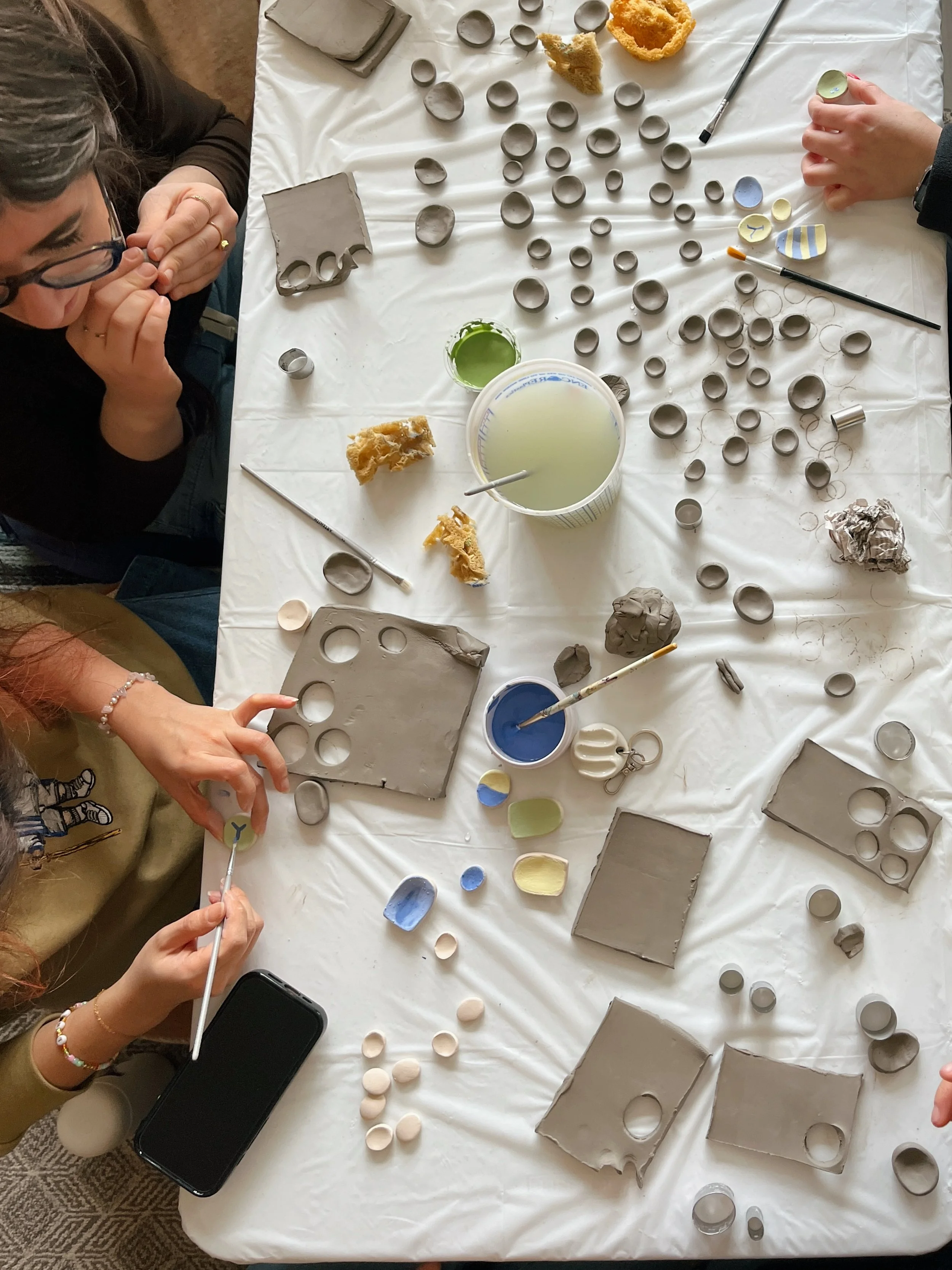 People working on a pottery project with clay, paint, and various tools on a white table.
