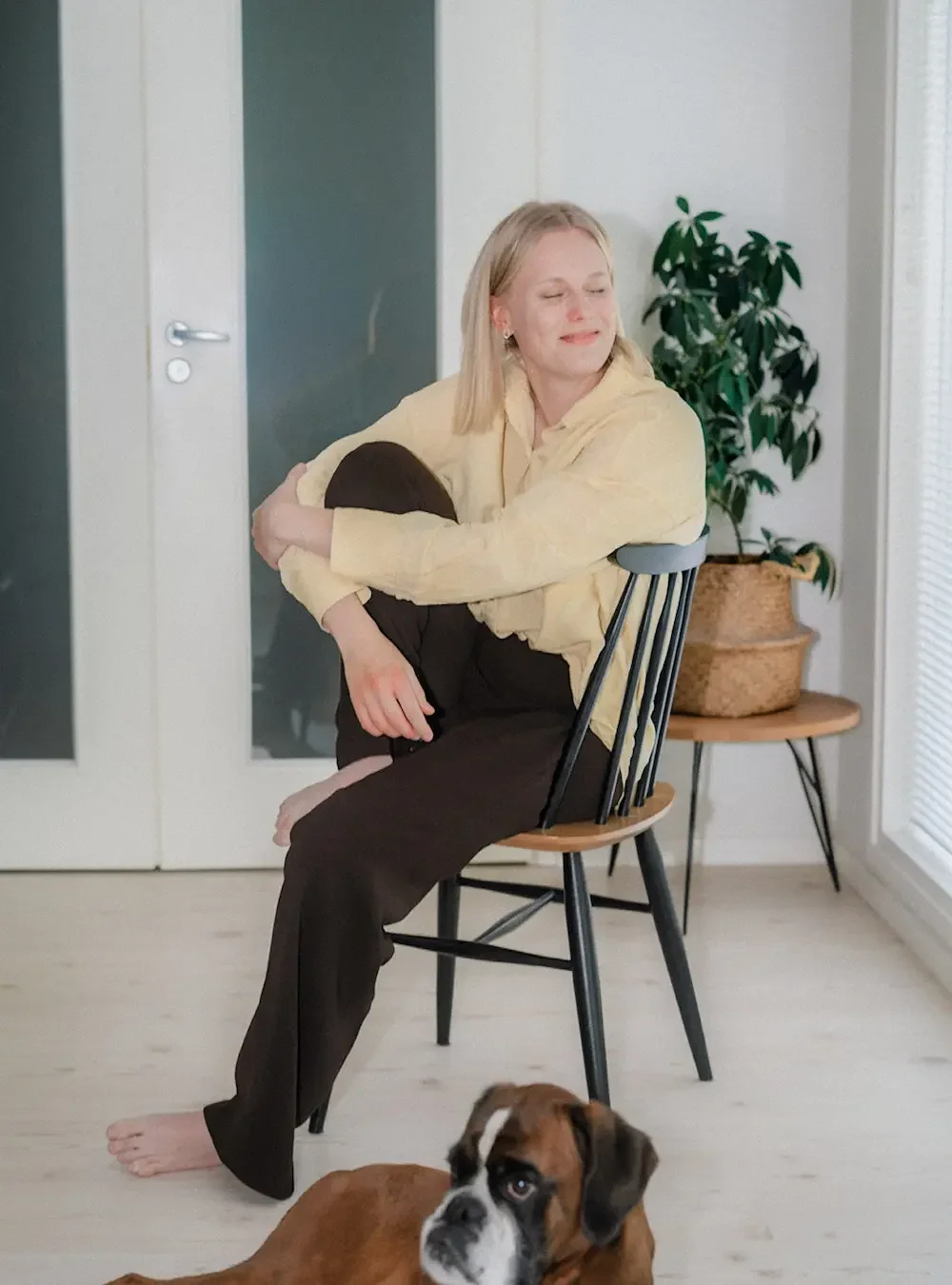 Nainen istuu tuolilla muotokuvassa, halailee polvea. Lattialla on koira. Taustalla ovet ja viherkasvi.