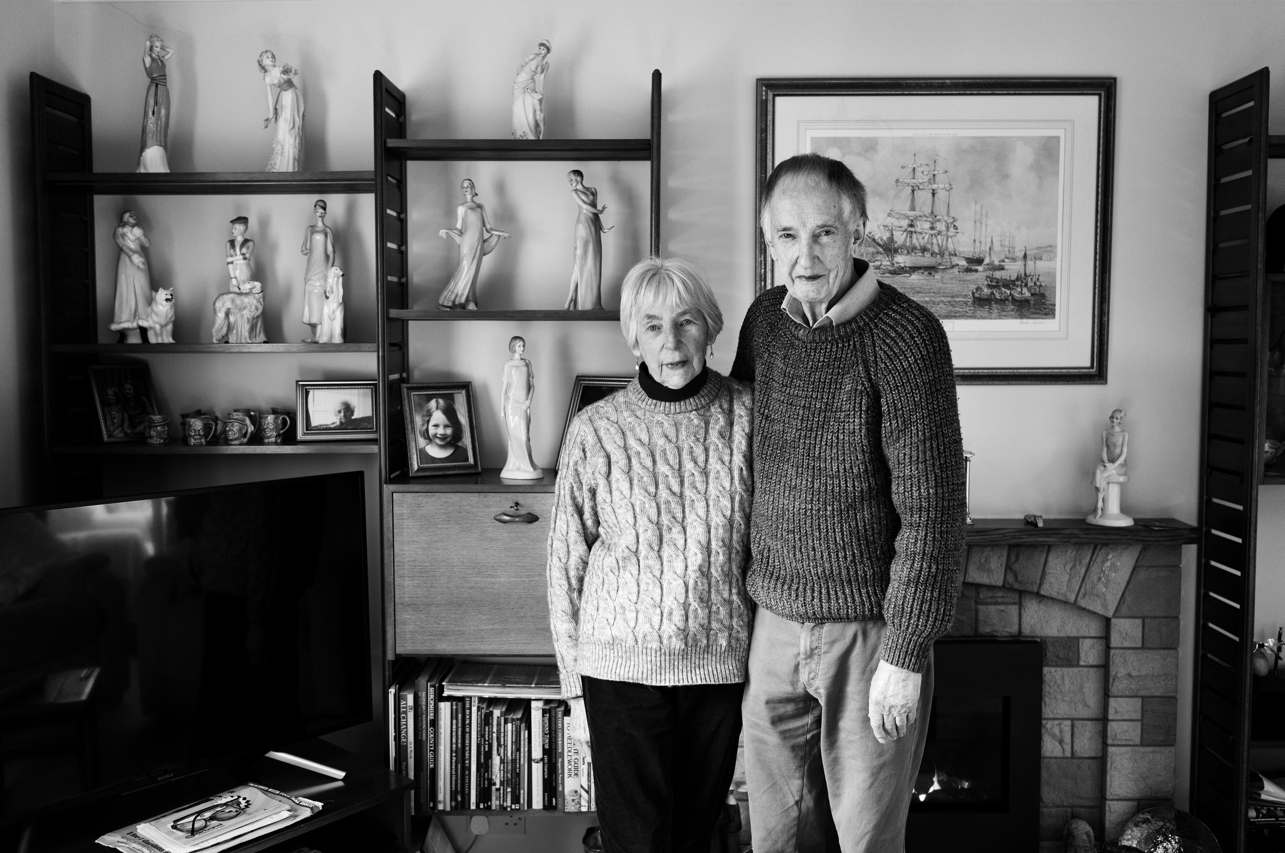 A portrait of an elderly couple (M/F) standing in what appears to be their living room. With a picture of some sailing ships behind them next to shelves filled with figurines.