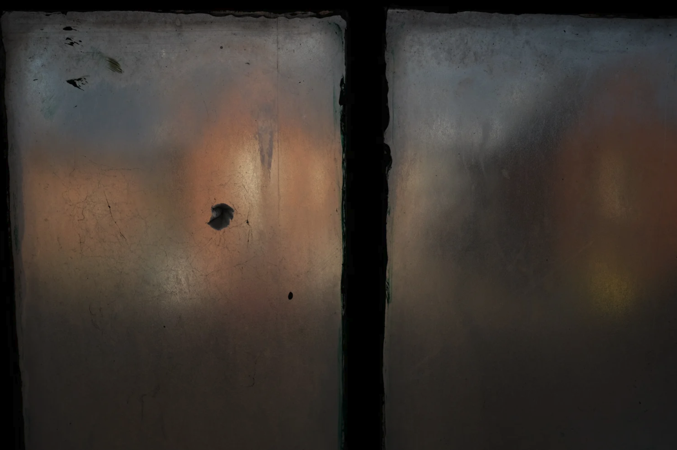 An image of a window looking outwards. The window is misty with condensation and so we can only see in a blurred way some red brick buildings lit by sunlight. The image is split in half by a part of the window frame/