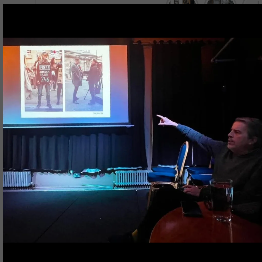 An of Patrick Willcocks ( a man of a certain age) sitting pointing towards one of his projected slides. The slide has 2 images; one of a man standing with a placard  '246 journalists killed'. The 2nd showing a TV interview of an RMT official.