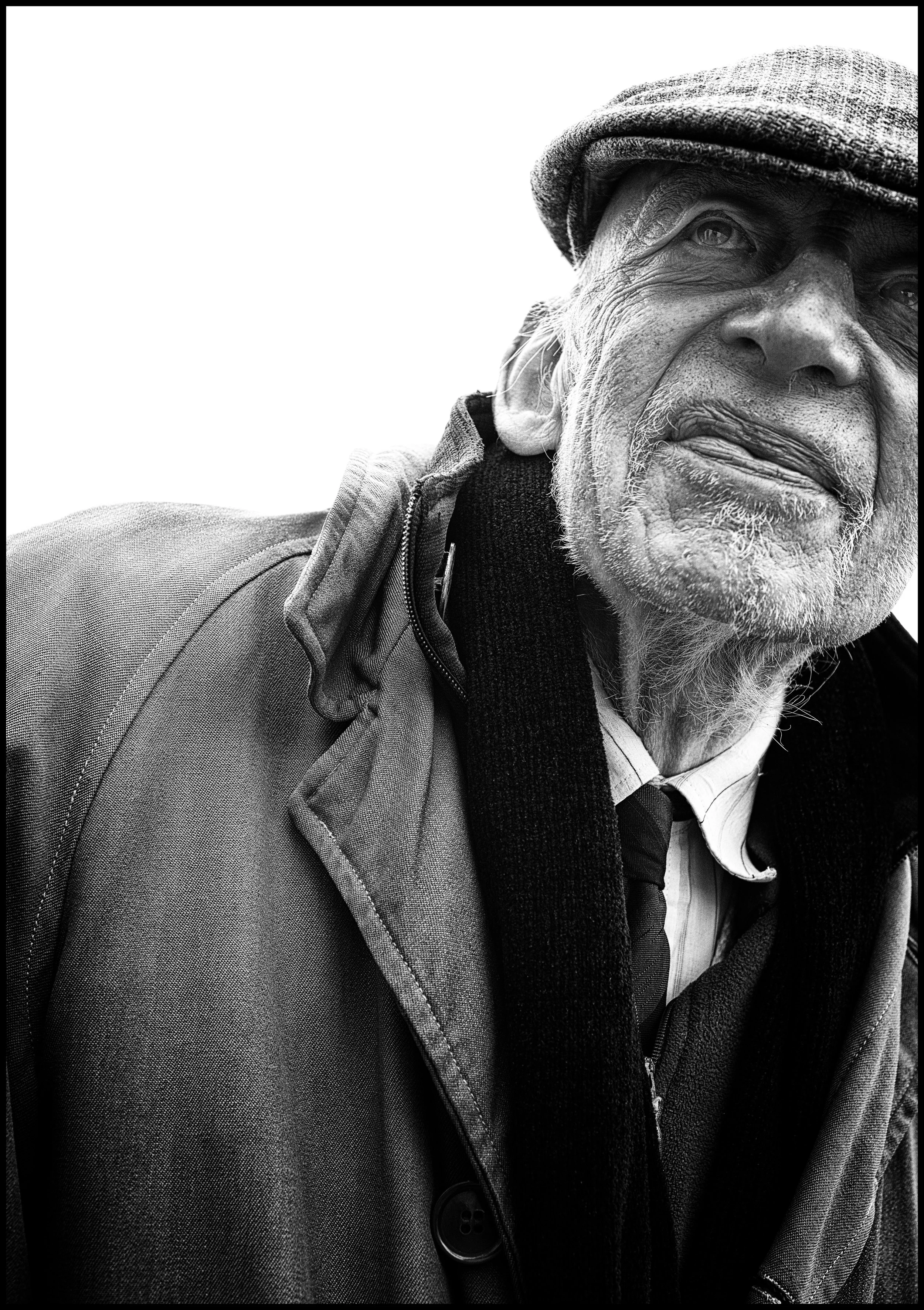A black and white portrait of an elderly white man dress in a raincoat with a hat/cap and black scarf. the image is looking up at him and is close up so not complete