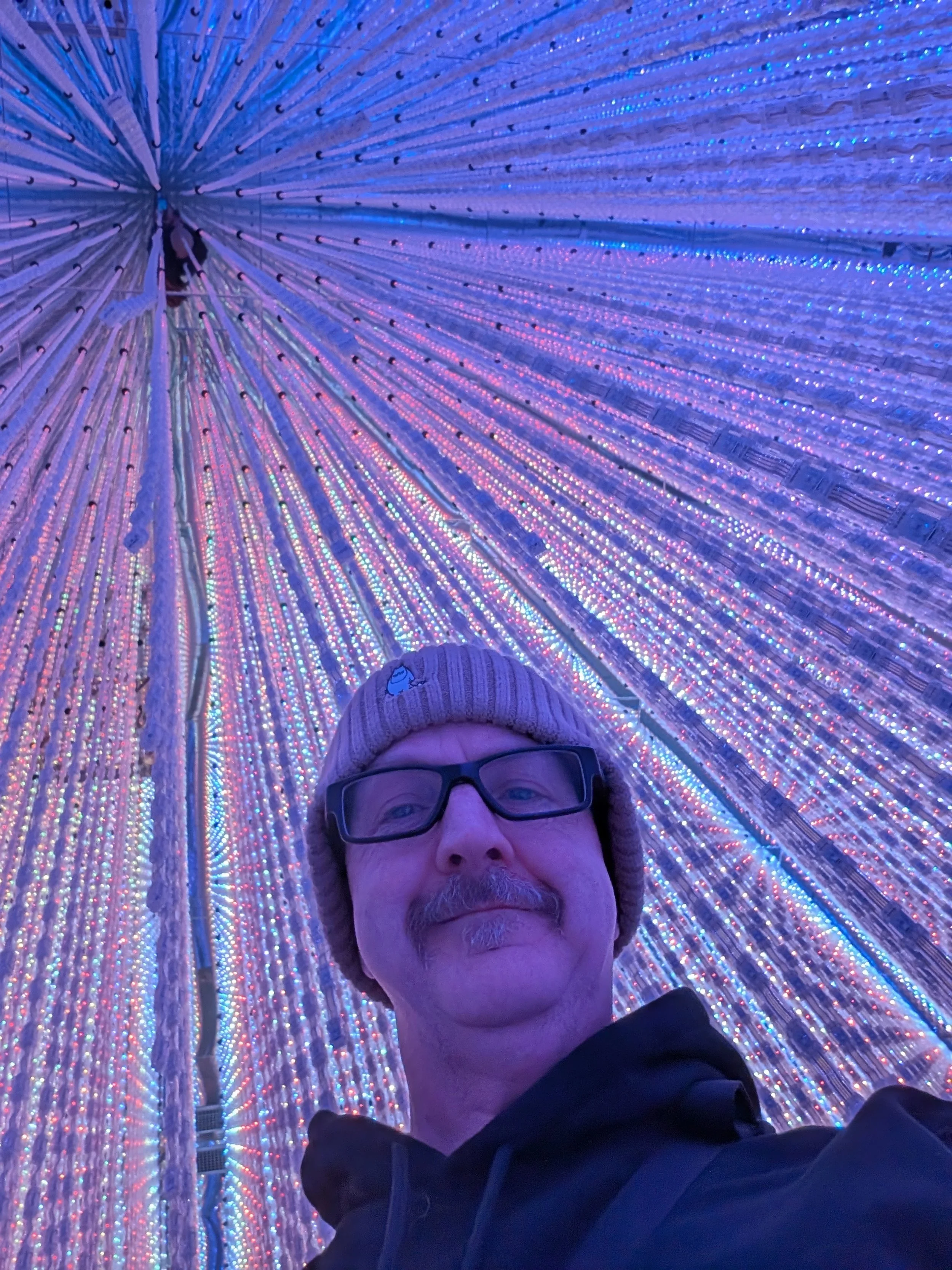 A picture of Neil Gibson (with black glasses and a beanie hat under a multi coloured awning.