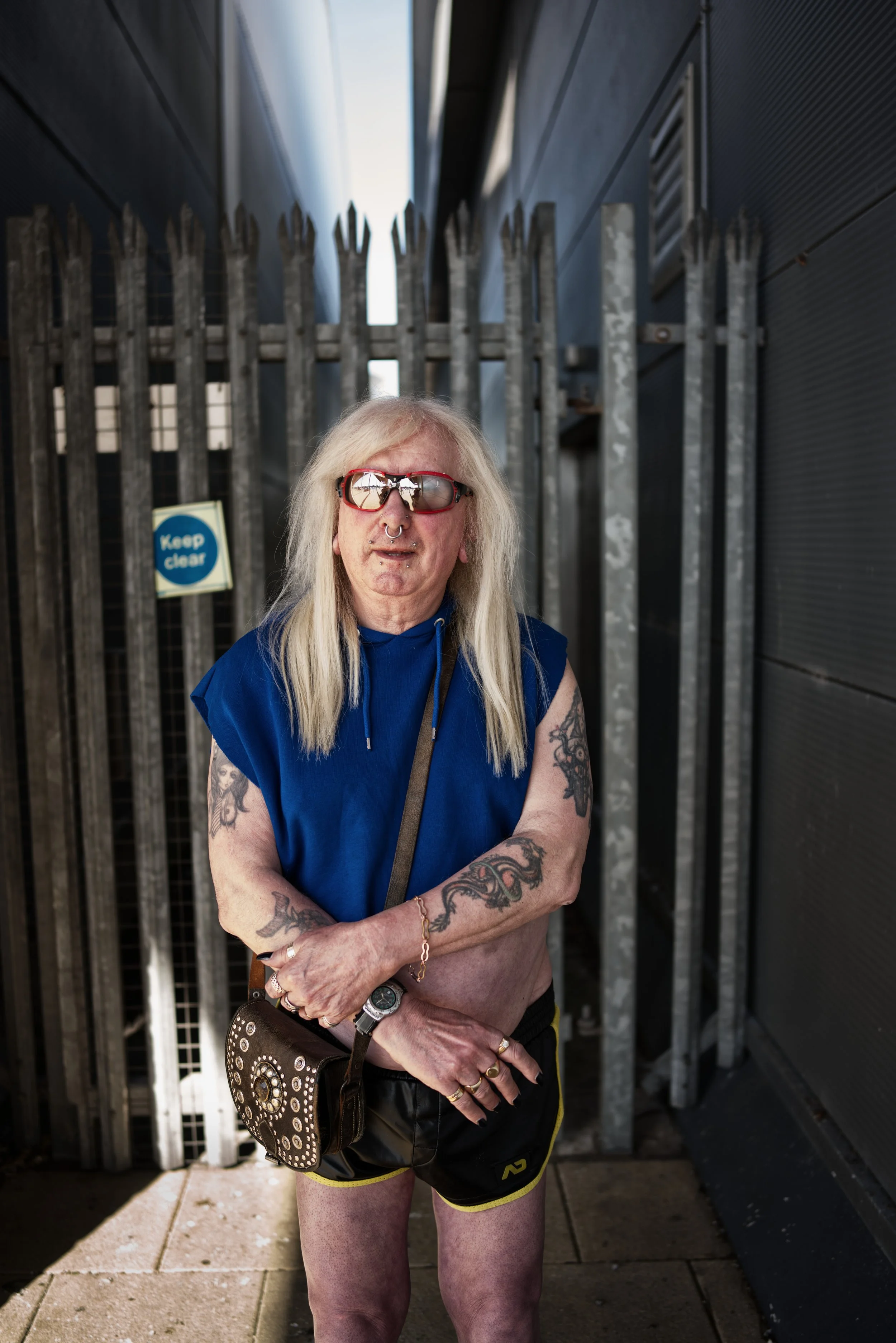 An image of a middle aged white man with long blond hair wearing shorts standing in front of a section of industrial fencing. He is wearing a blue top and sunglasses.