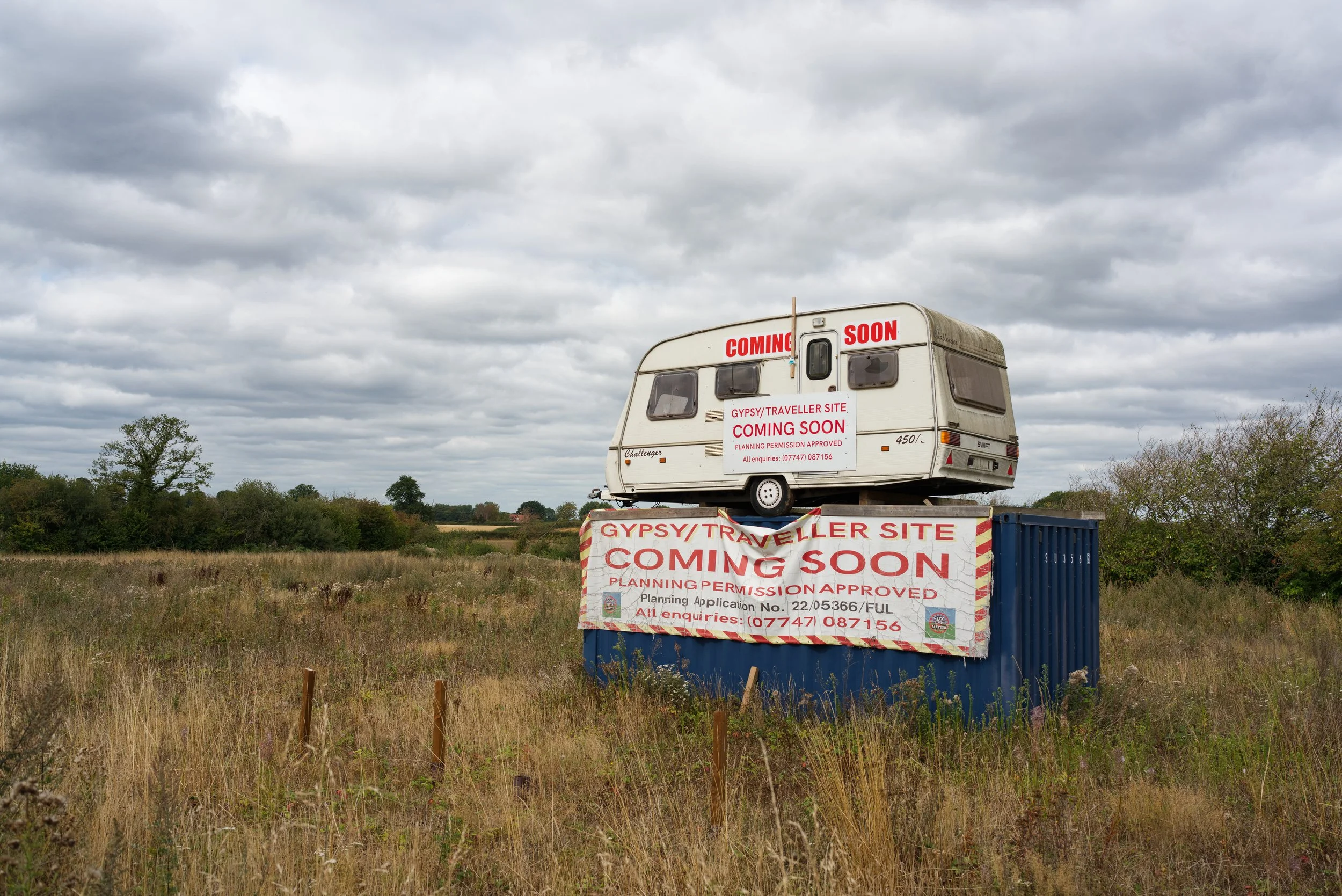 A colour landscape image of a caravan bizarrely parked sitting on a shipping container in the middle of a field. Both are draped with banners highlighting the imminent arrival of a 'gypsy traveller site. Seemingly as a protest.