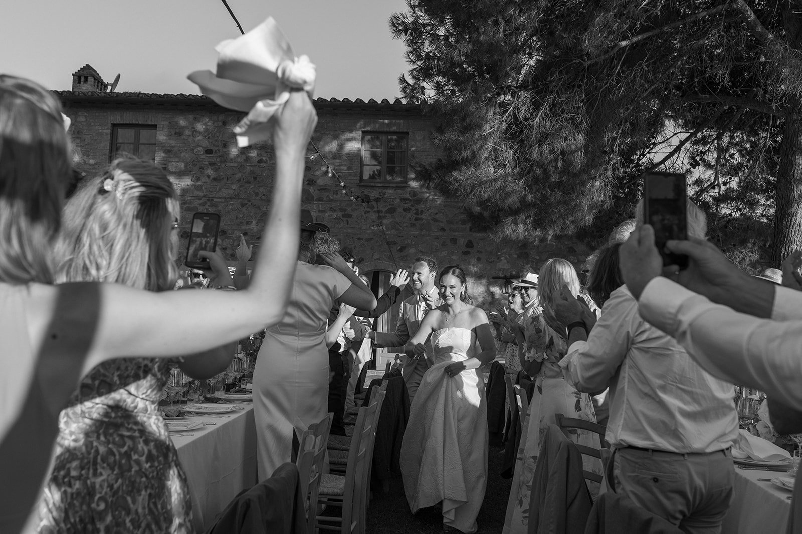 People celebrating at an outdoor wedding reception, with a bride and groom walking through the crowd, guests taking photos and high-fiving, tables and chairs, trees and a stone building in the background.