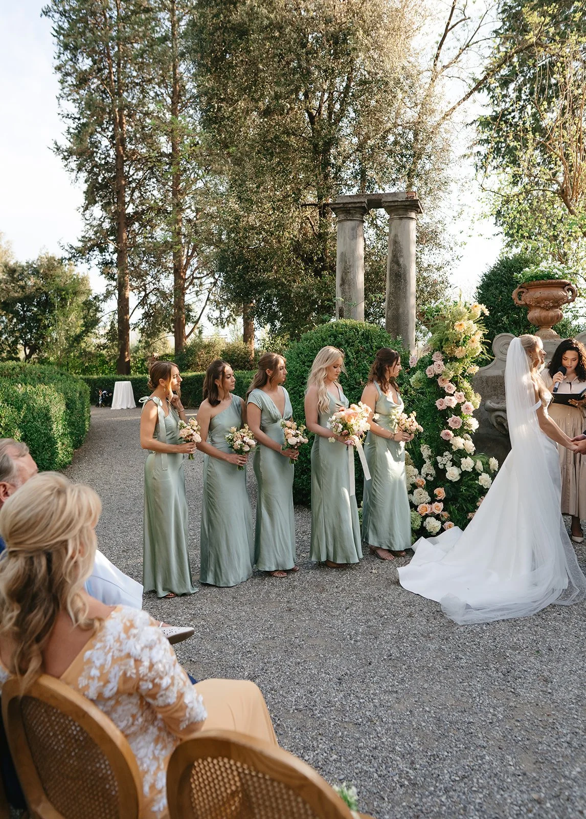 A wedding ceremony outdoors with bridesmaids in matching light green dresses standing in a row holding bouquets, and the bride in a white dress with a veil and a bouquet, standing in front of a floral arrangement and ancient ruined columns on a grave