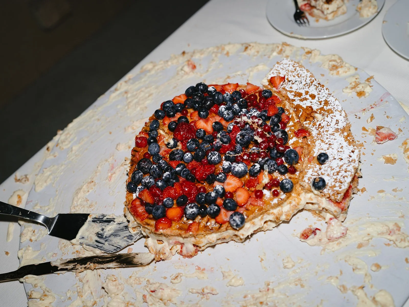 A partially eaten fruit tart topped with mixed berries, including strawberries, blueberries, and red currants, on a white surface with remnants of cream and pastry crust.