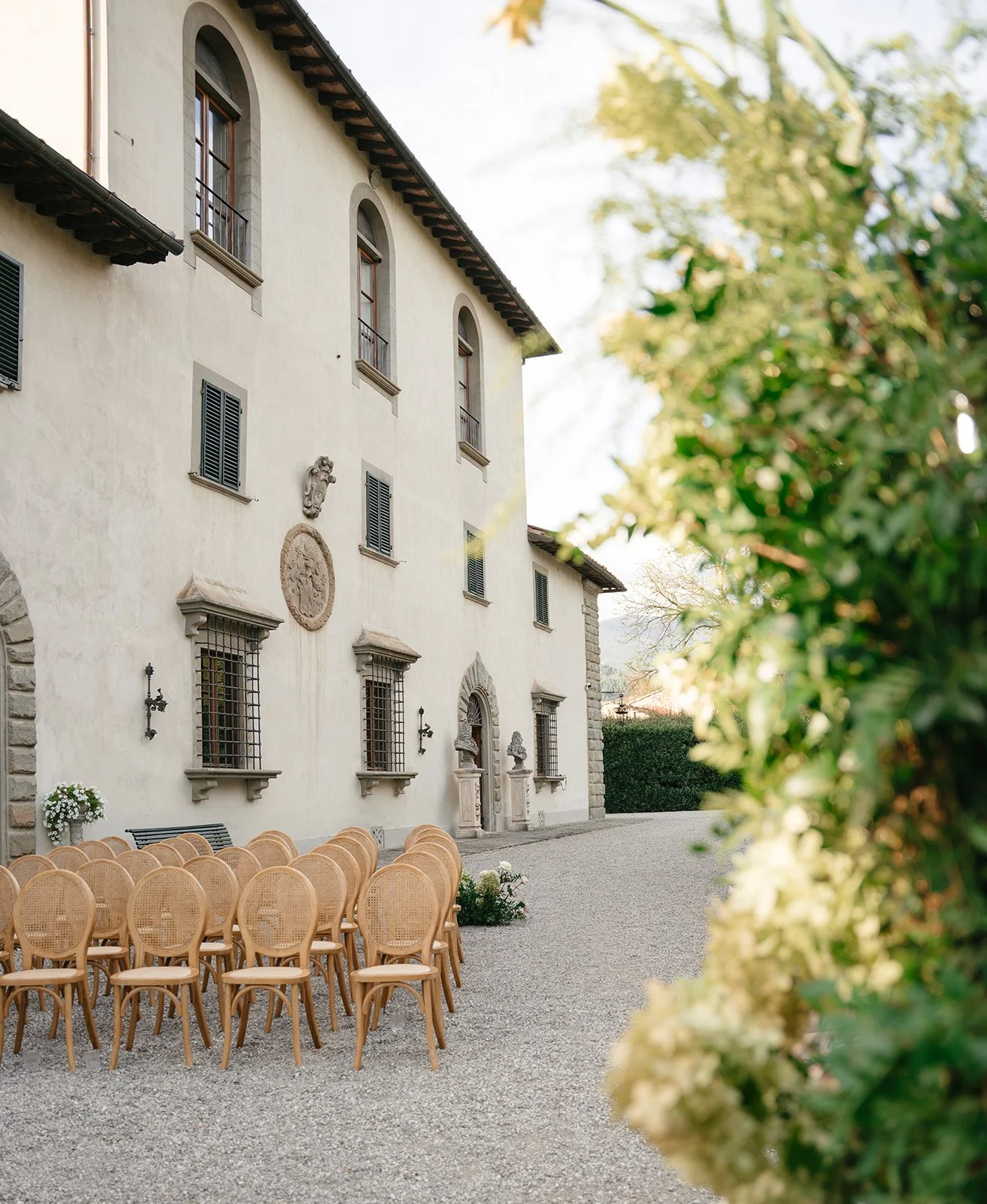 Outdoor courtyard with beige chairs arranged in rows, decorated with flowers, in front of an old European-style building with arched windows, stone decorations, and a gravel ground.
