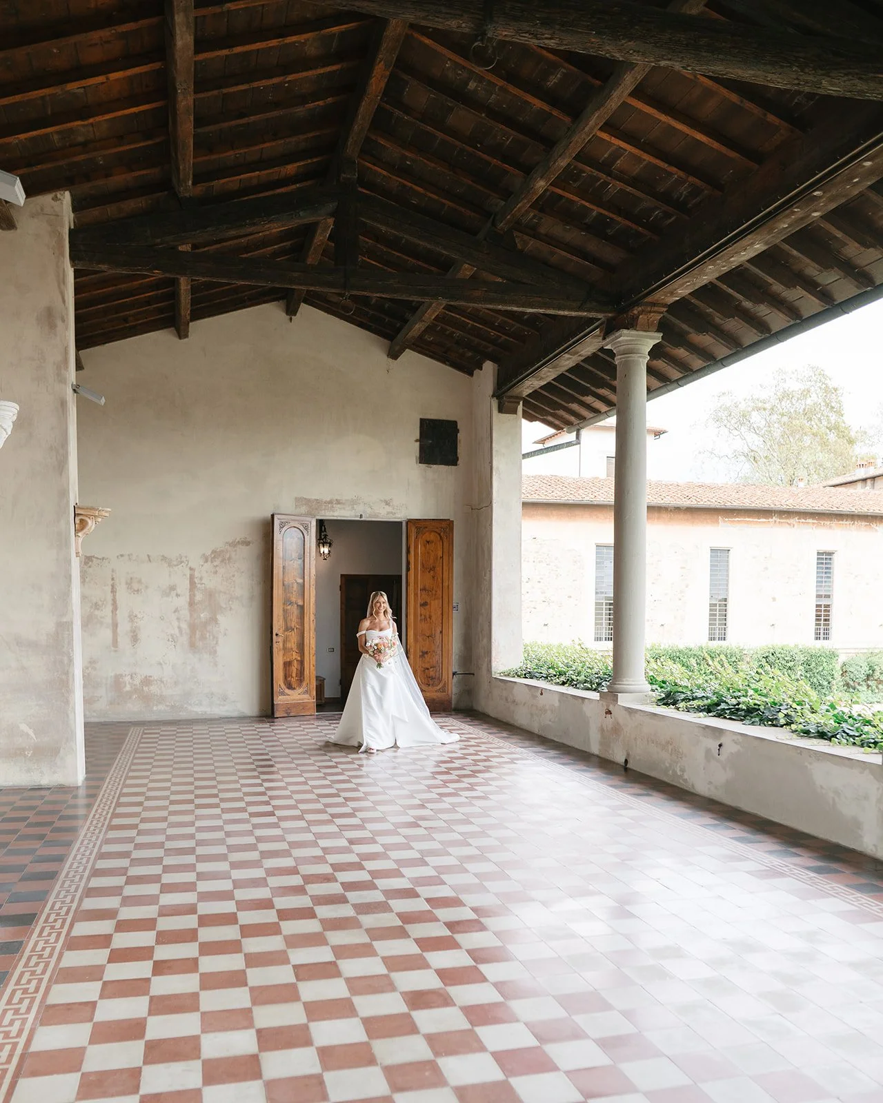 A bride in a white wedding dress holding a bouquet standing in an open area with high wooden ceiling and checkered floor, doorway behind her.