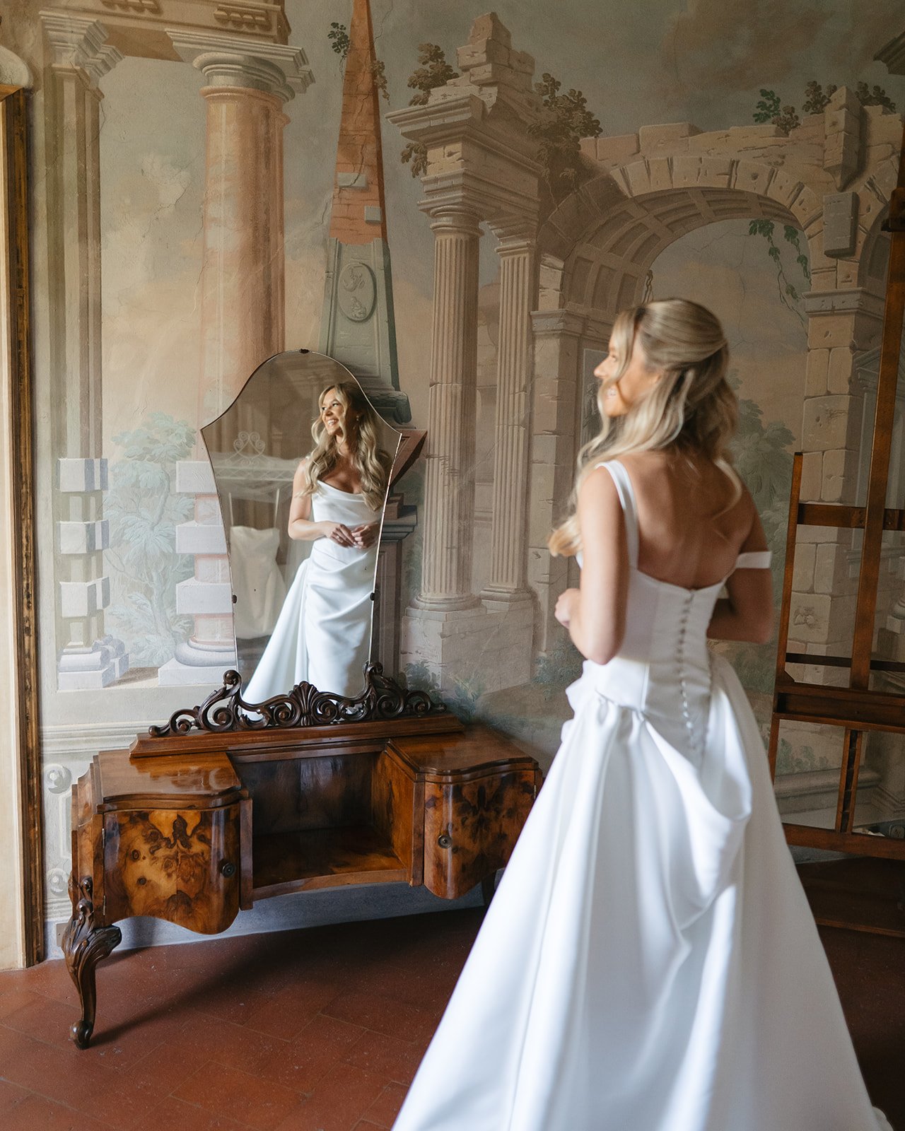A woman in a white wedding dress is looking at herself in a decorative oval mirror on a vintage wooden dressing table. The setting is a room with a mural of ancient ruins, including columns and arches.