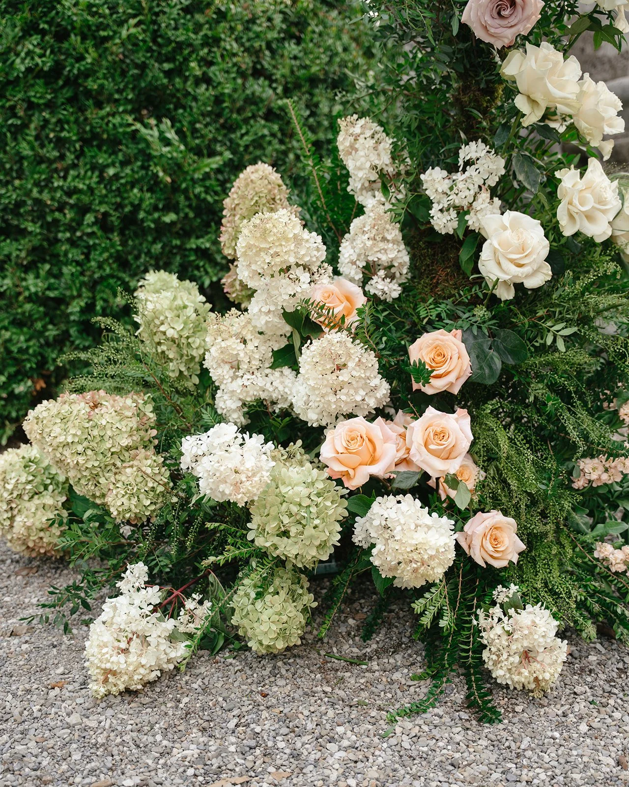 A floral arrangement of white hydrangeas, pale pink roses, and white roses with green leaves on a gravel ground against a green shrub background.