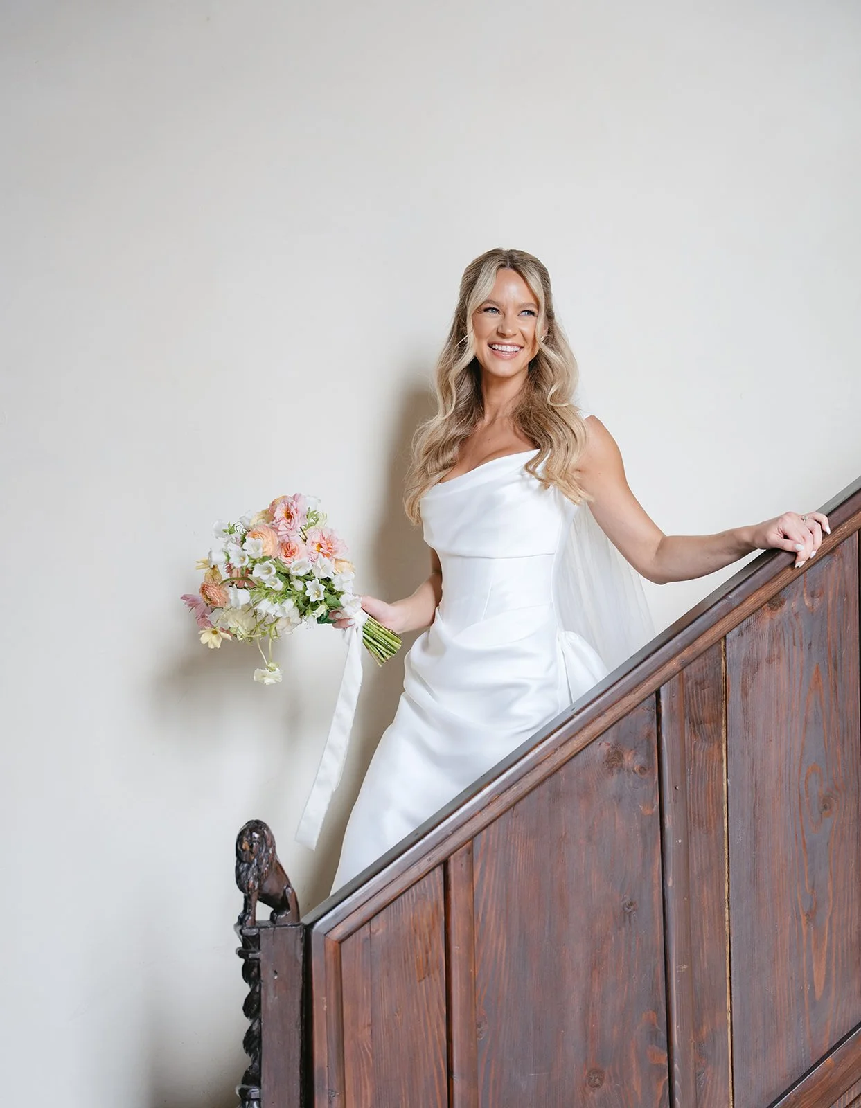 A woman in a white wedding dress holding a bouquet of pink and white flowers, standing on a staircase with a wooden railing, smiling.