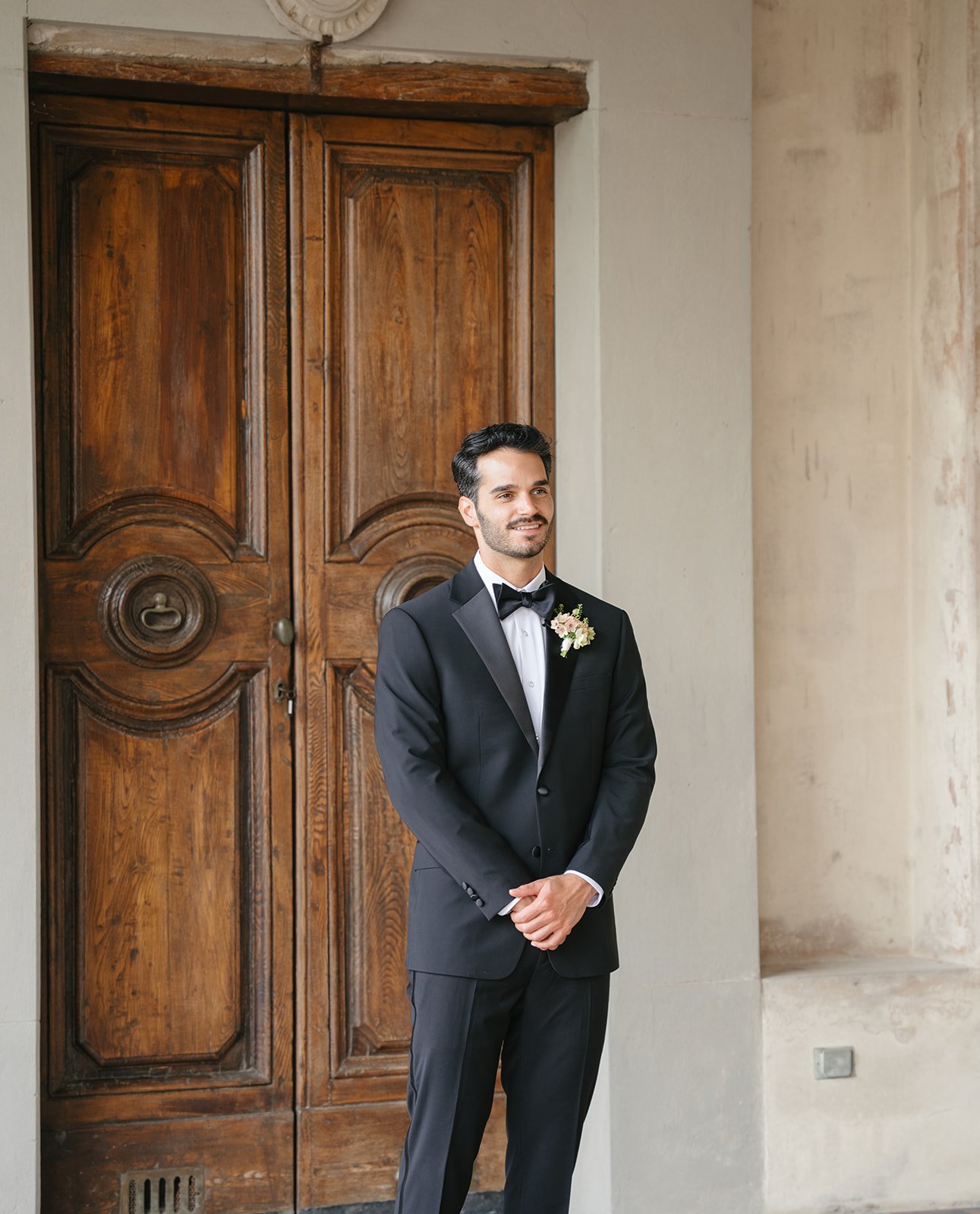 A groom in a black tuxedo with a bow tie and a boutonniere, standing with hands clasped, smiling in front of a large wooden door and a stone wall.
