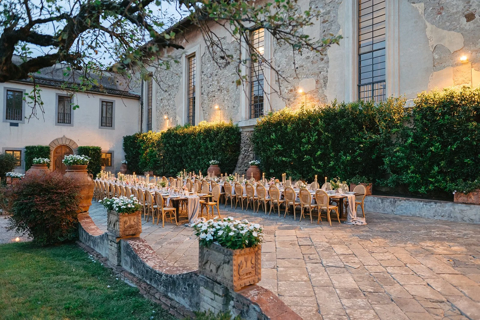 Outdoor dining setup with long banquet table, candles, and flower arrangements, in a courtyard next to a historic stone building, during evening.