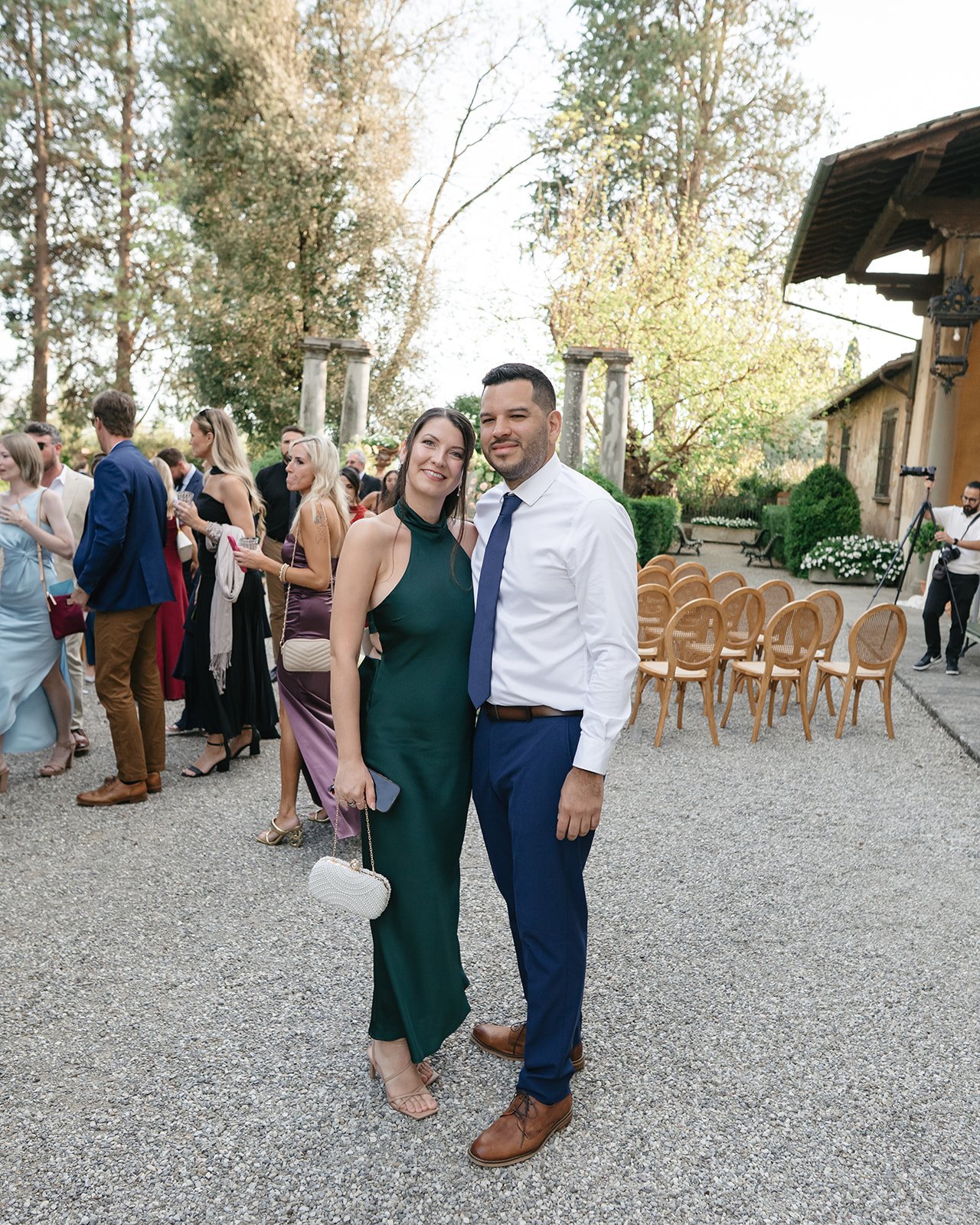 A man and woman dressed nicely pose together at an outdoor event, smiling, with guests and trees in the background.