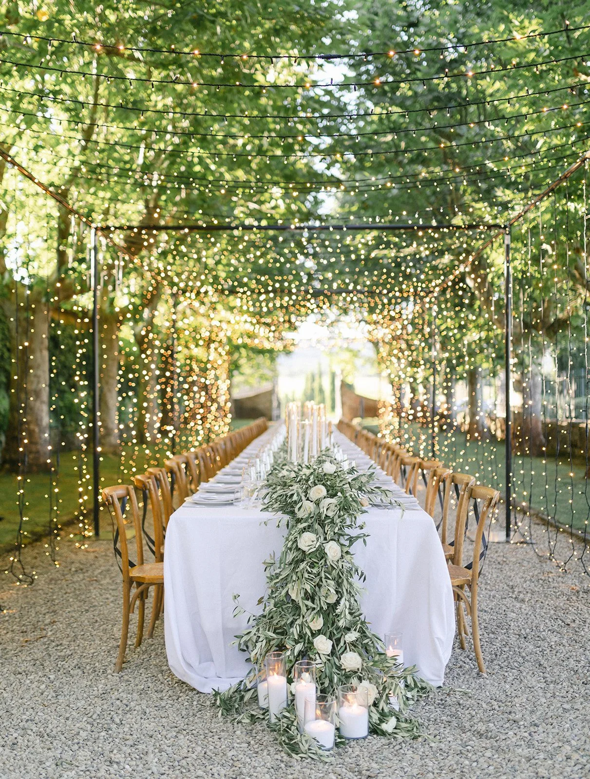 Outdoor dining table decorated with white tablecloth, floral centerpiece of white roses and greenery, surrounded by wooden chairs, under string lights and twinkling fairy lights in a green, leafy setting.