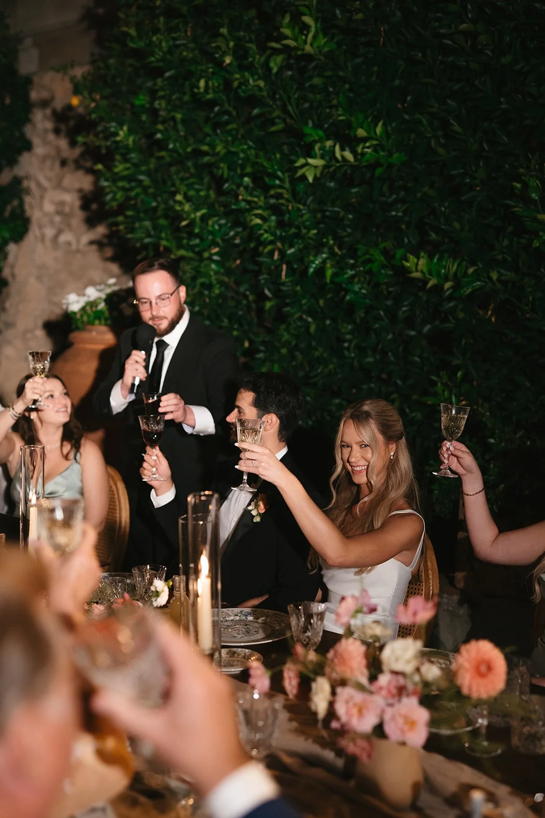 People celebrating at a wedding reception, toast with glasses, a woman in a white dress smiling, and a man in a tuxedo, with green foliage in the background.