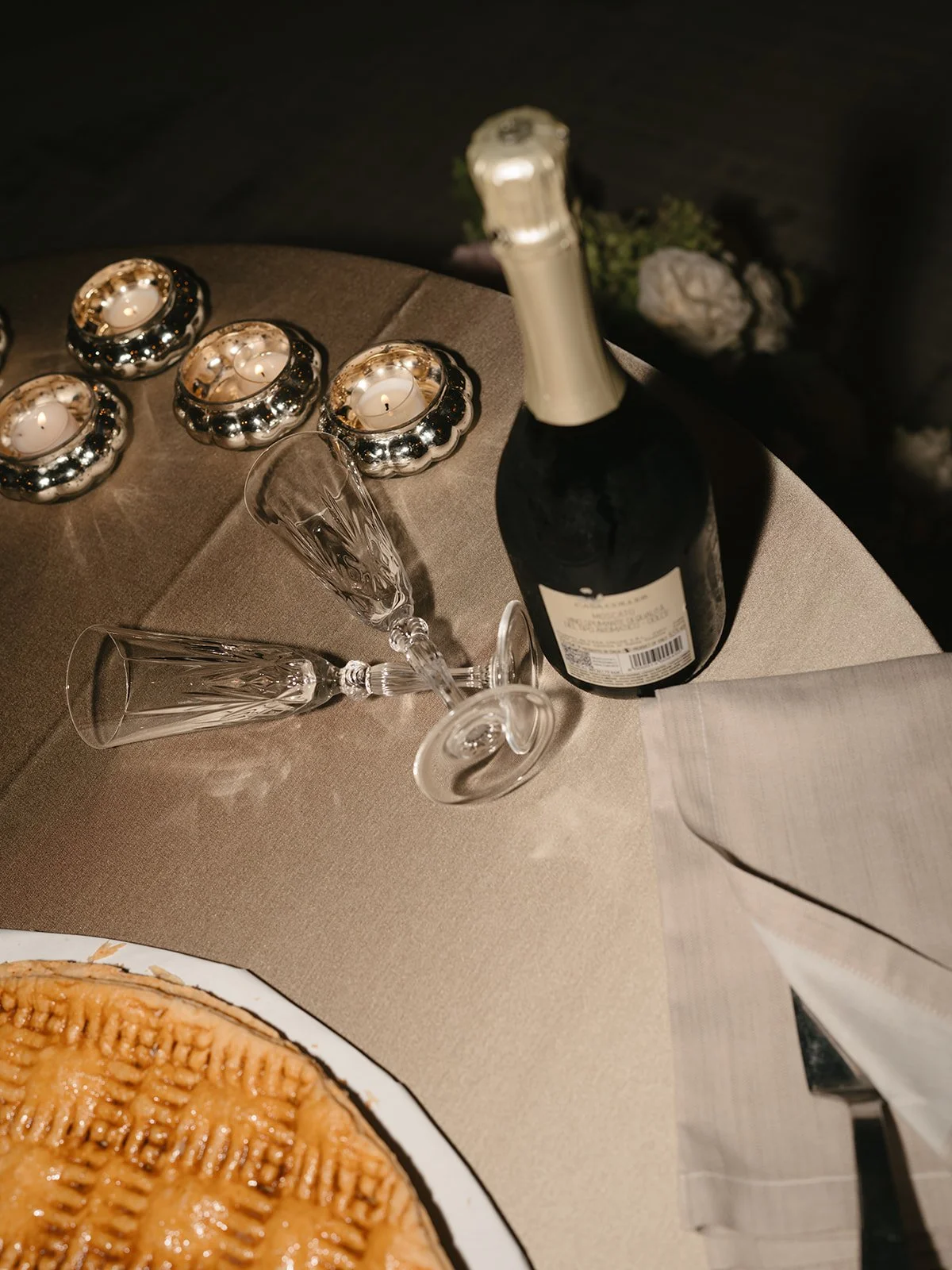 Table setting with a bottle of champagne, two champagne flutes, candles in decorative holders, and part of a pie on a plate.