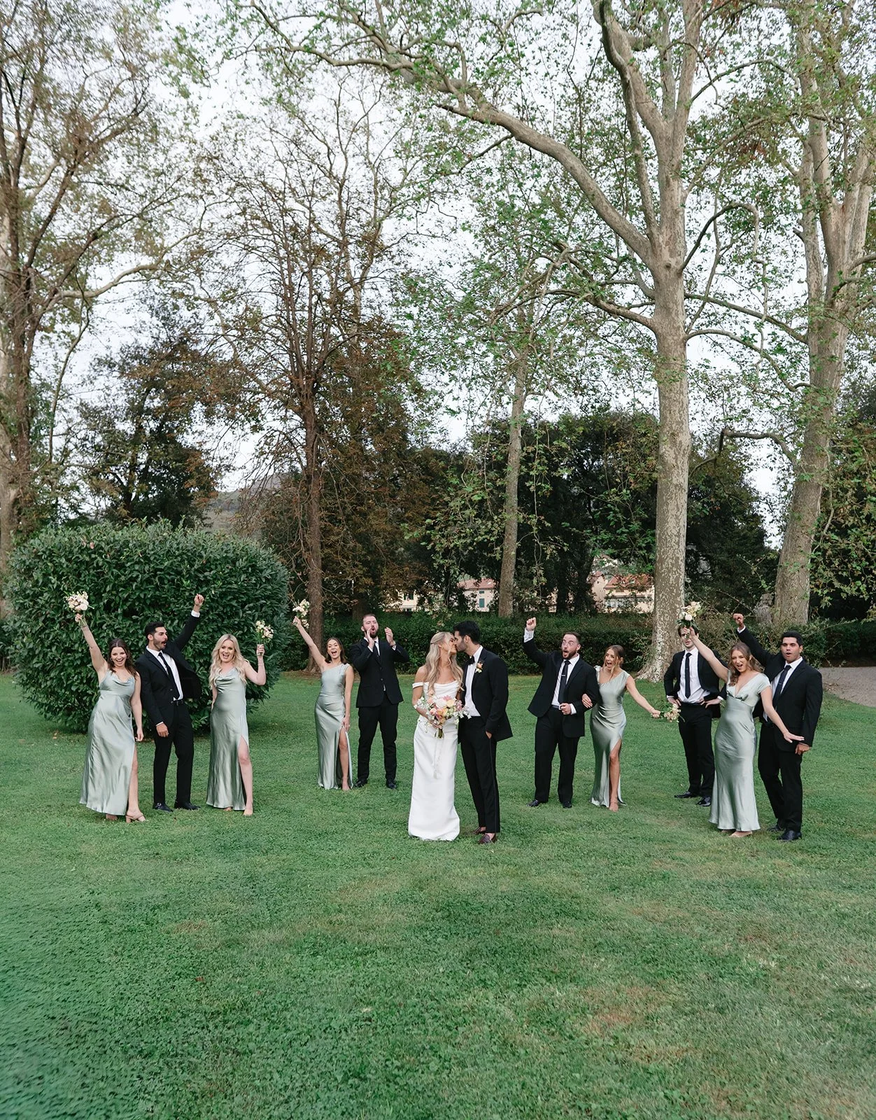 A wedding party of ten, including the bride and groom, celebrates outdoors on grass with trees in the background. The women wear silver dresses, and the men wear black suits. Some hold bouquets and raise their fists in celebration.