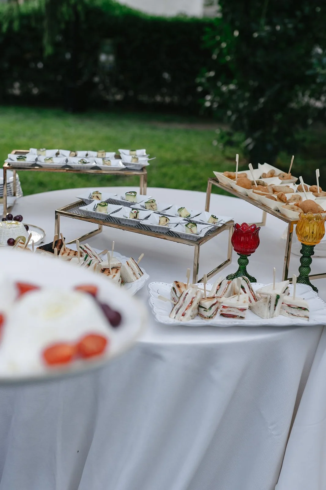 A table with assorted finger foods including sandwiches, cucumber roll-ups, and pastries, set outdoors with a white tablecloth and decorative glassware.