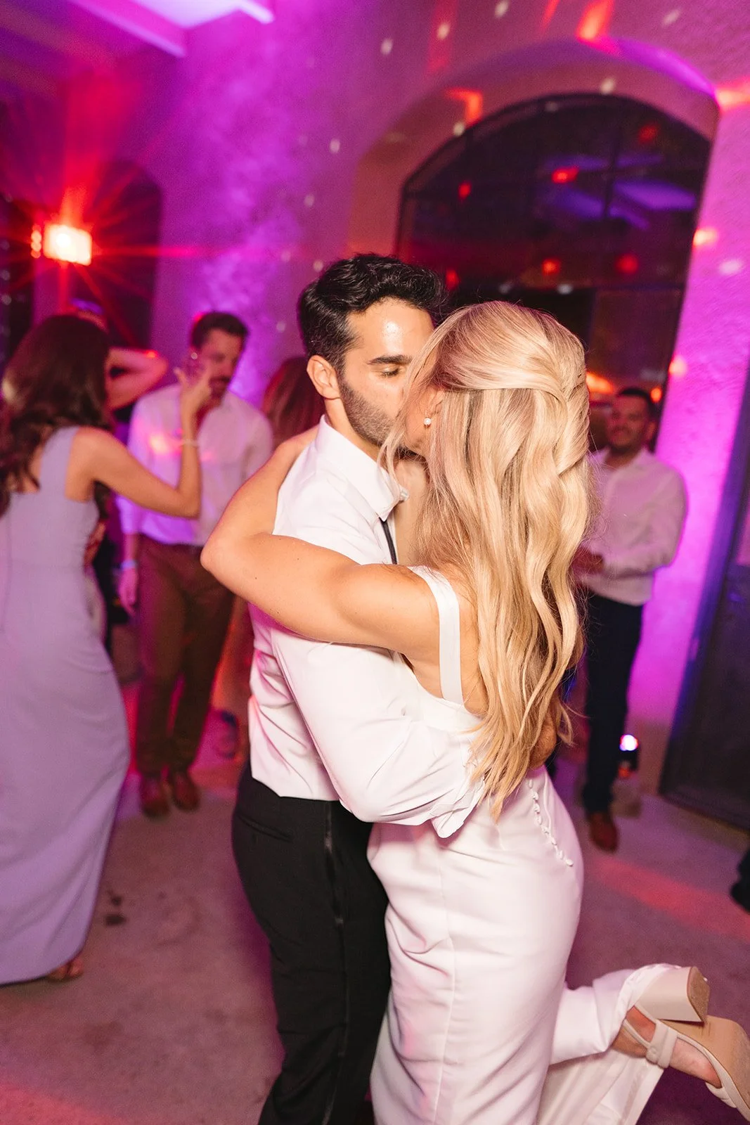 Couple dancing closely at a party, with colorful purple and pink lighting and other guests in the background.