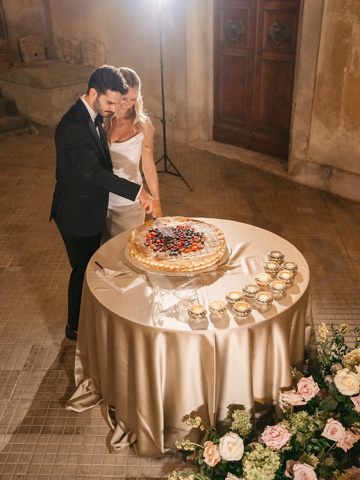 A bride and groom cutting a wedding cake at a wedding reception with candles and flowers on the table