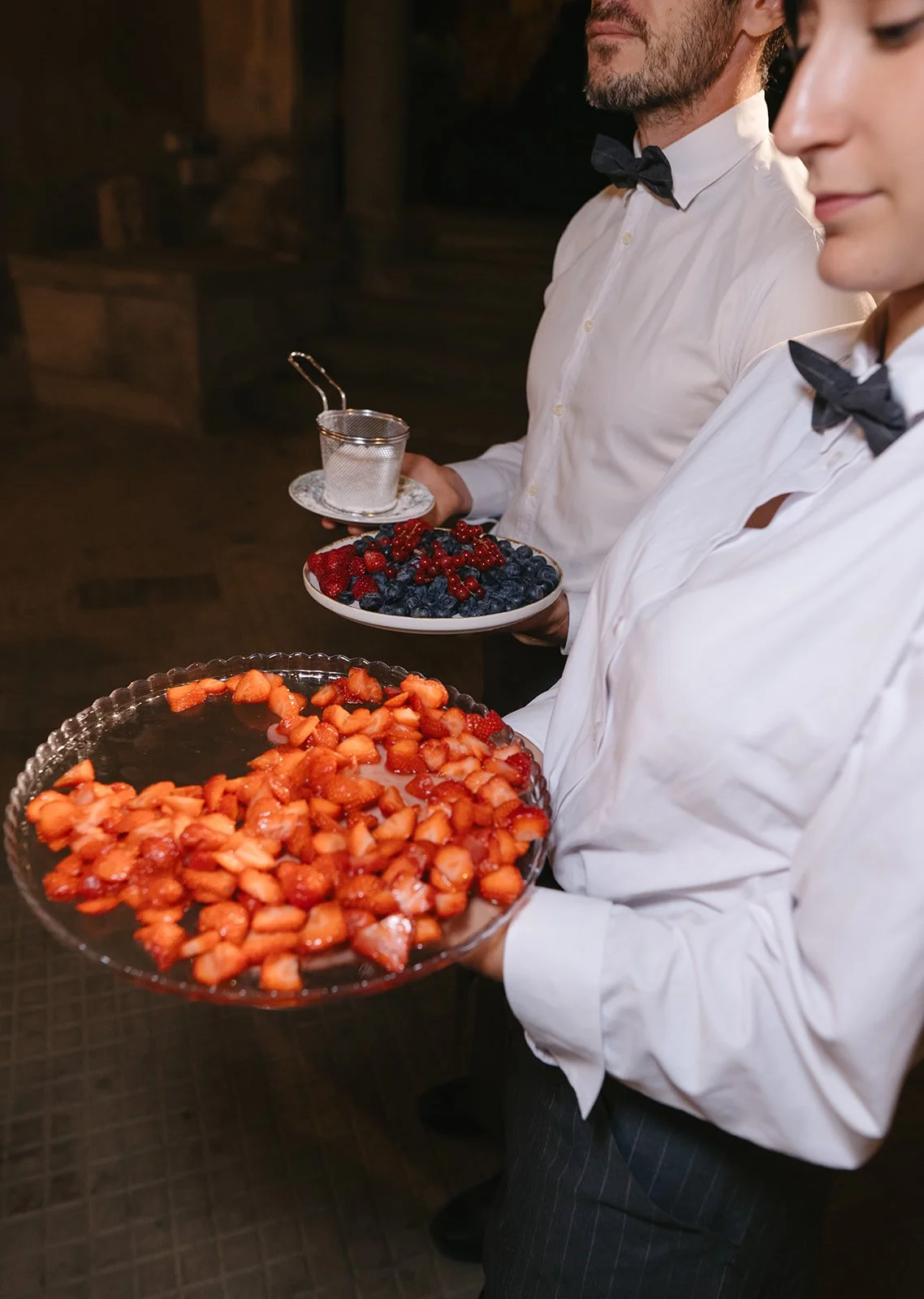 Two servers dressed in white shirts with black bow ties holding trays of fresh strawberries and mixed berries at a formal event.