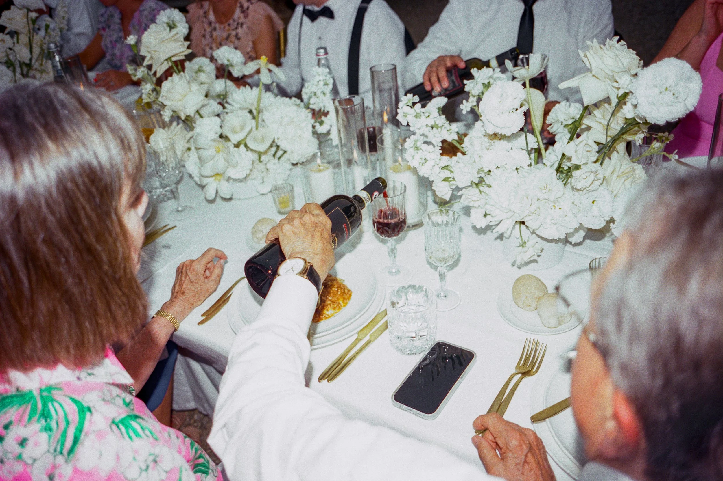 People sitting around a table with white floral arrangements, pouring wine, and enjoying a meal at a formal event or celebration.