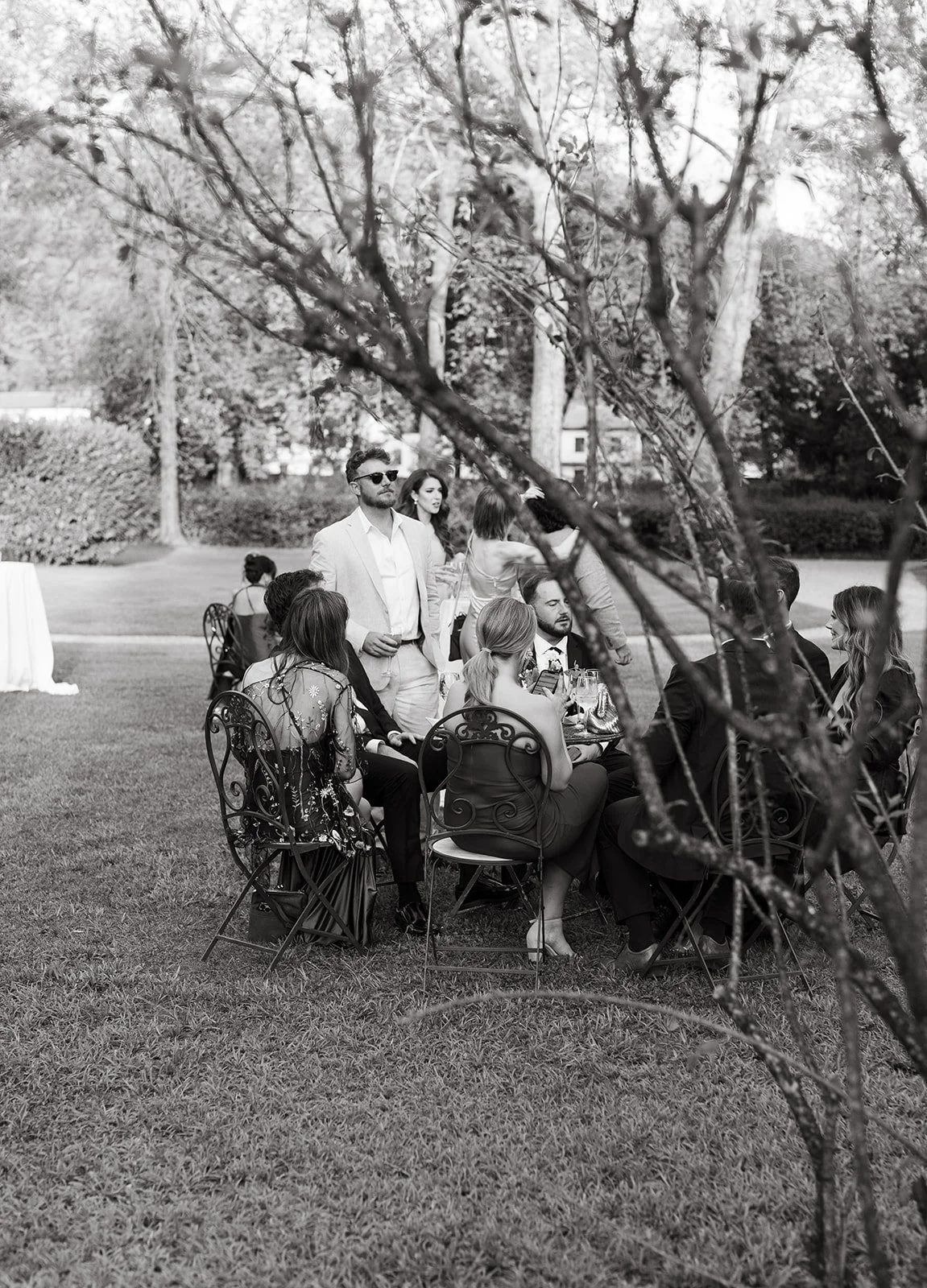 A group of people dining outdoors at a formal event, seen through tree branches. The guests are seated around a table with drinks, and some standing, dressed in business or formal attire.