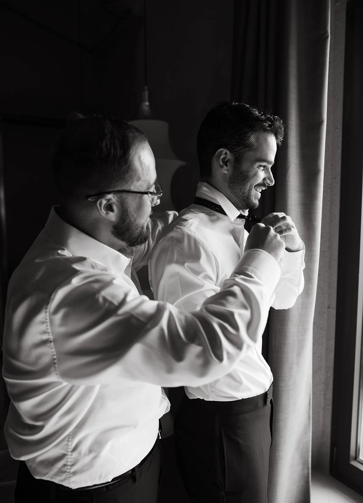Two men in formal attire adjusting a tie and preparing for a formal event near a window.