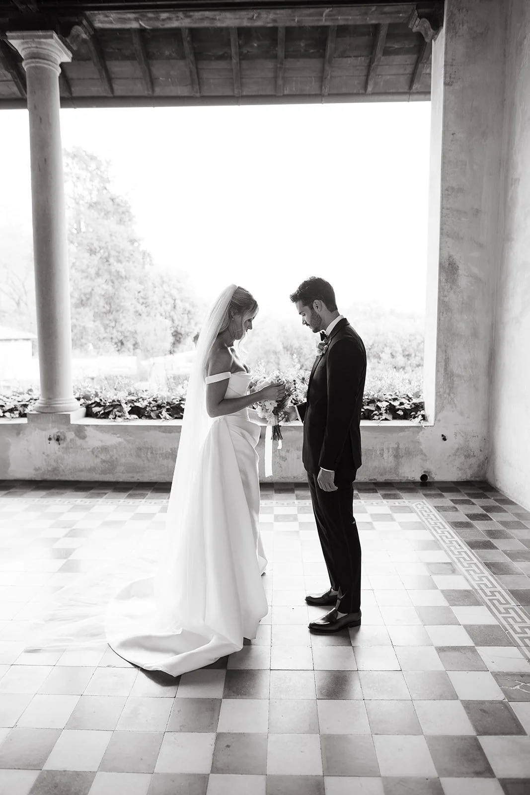 Black and white photo of a bride and groom standing close together, looking at each other, with the bride holding a bouquet of flowers. They are in a room with tiled floor and an open window in the background, framed by two columns, with trees visibl