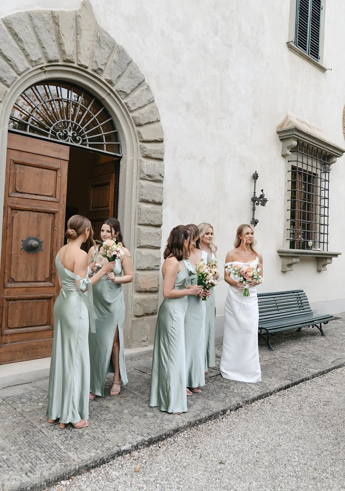 Group of bridesmaids standing outside a building with large wooden doors, holding bouquets, preparing for a wedding.
