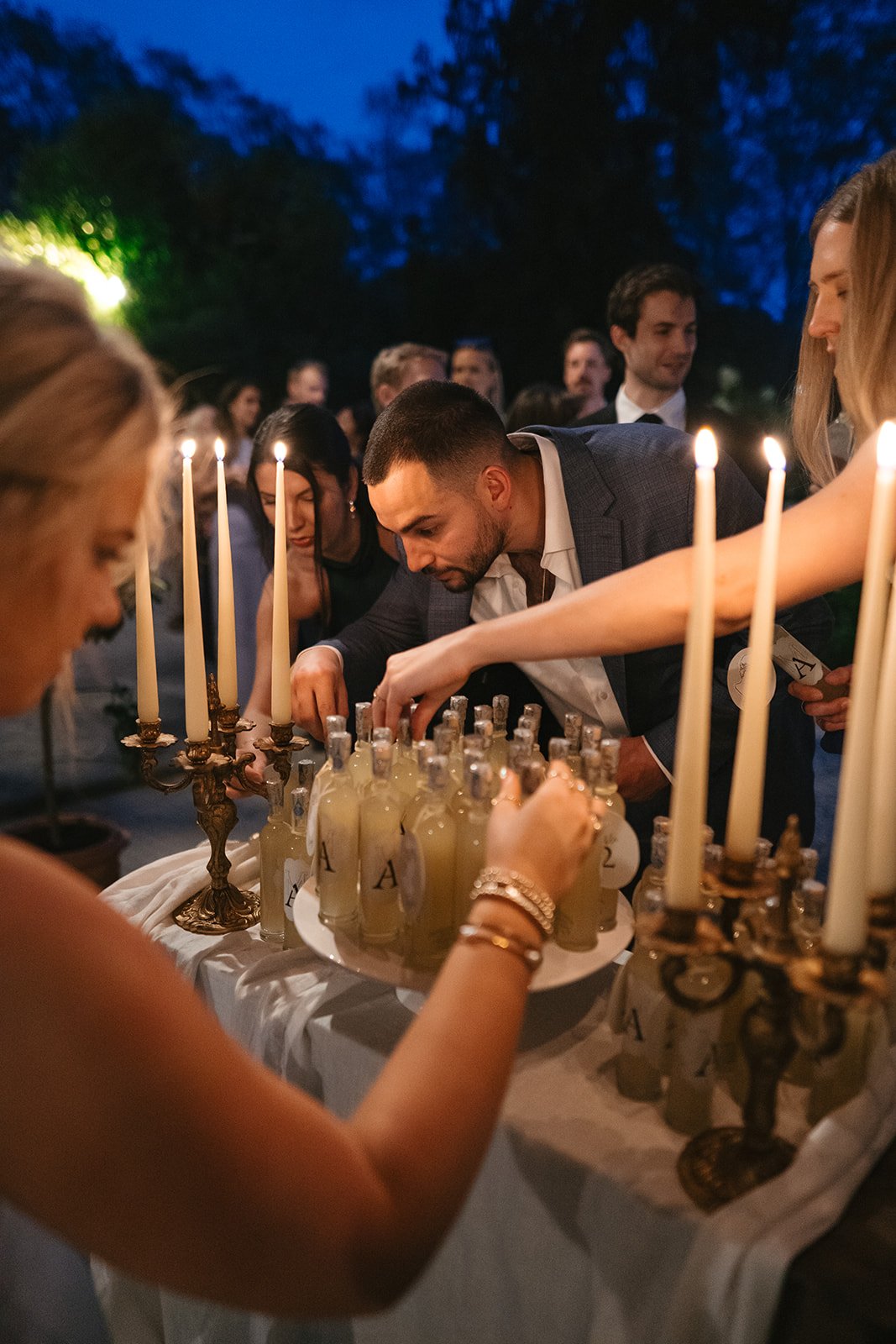 People gather around a table with bottles and lit candles during an outdoor evening event or celebration.
