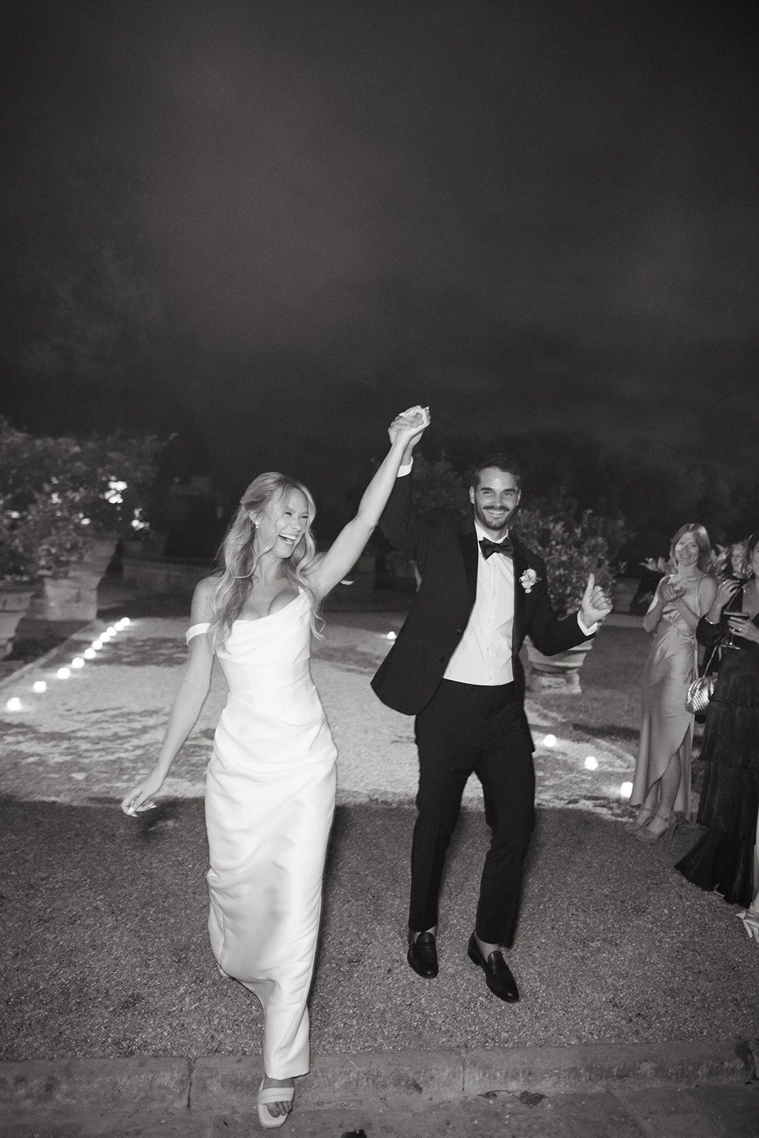 A black-and-white photo of a newlywed couple celebrating at night. The bride, in a strapless wedding gown, has her hand raised and is smiling. The groom, in a tuxedo, is holding her hand and giving a thumbs-up. Guests are visible in the background.
