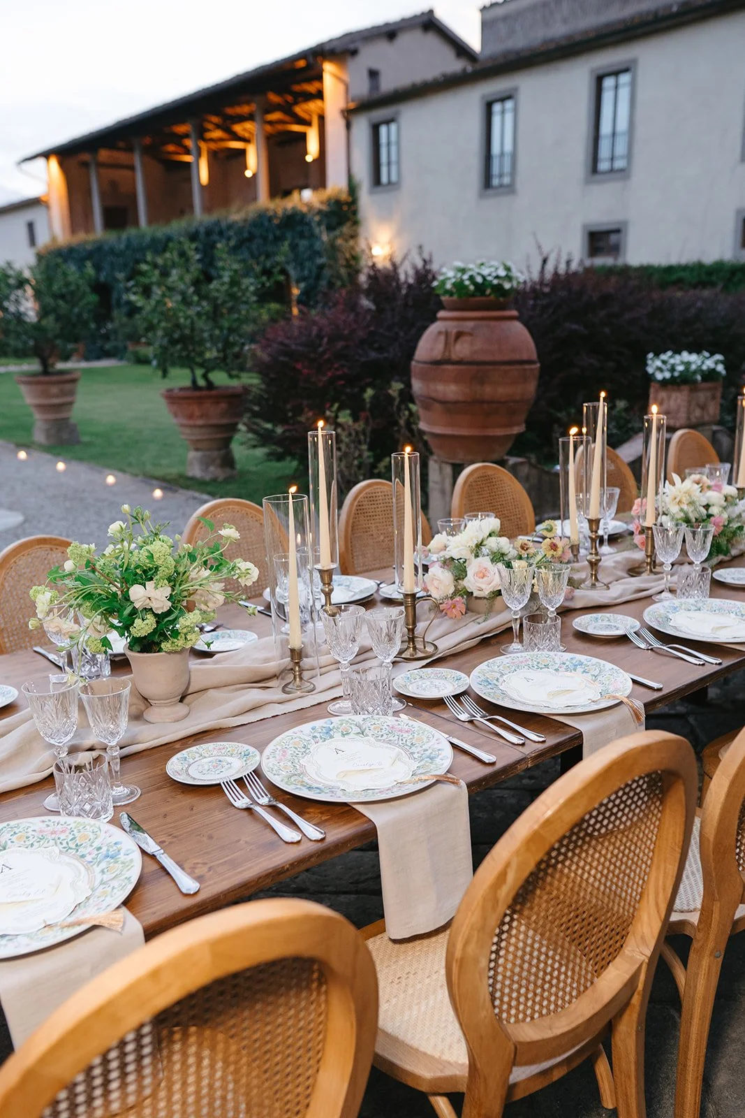 An outdoor dining table set for a formal event with floral centerpieces, candle holders, fine china, and silverware, on a patio with a house and garden in the background.