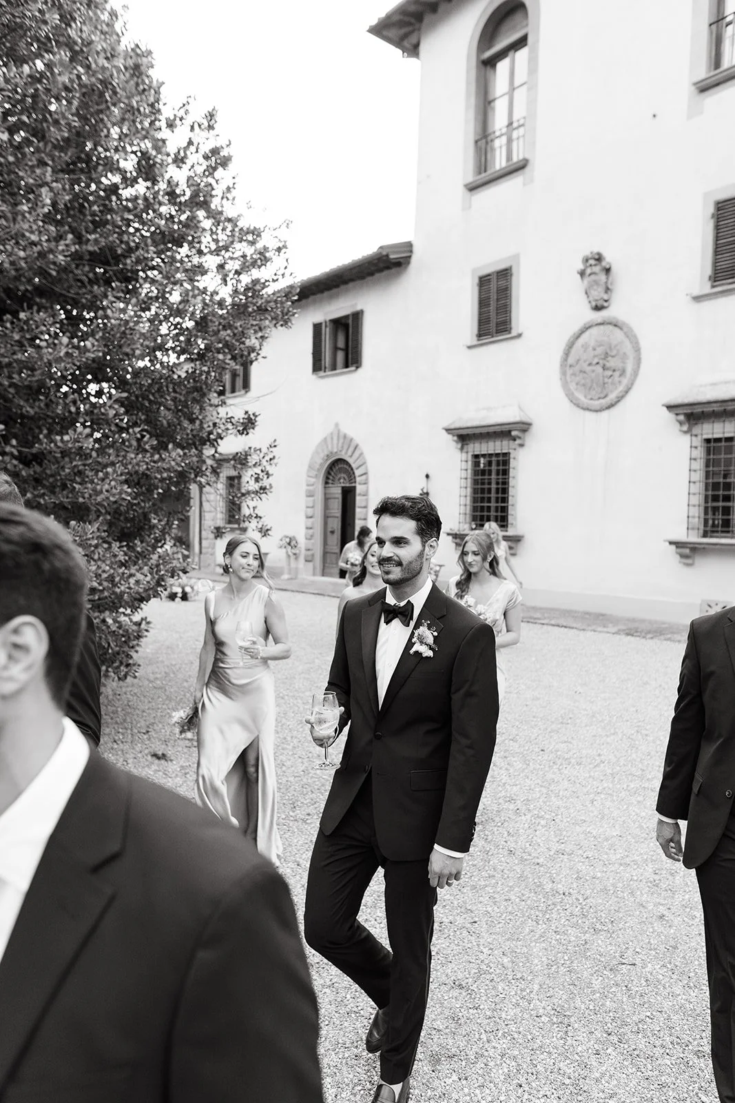 A groom in a tuxedo walking outside with guests, holding a wine glass, in front of an elegant villa with windows and decorative emblems.